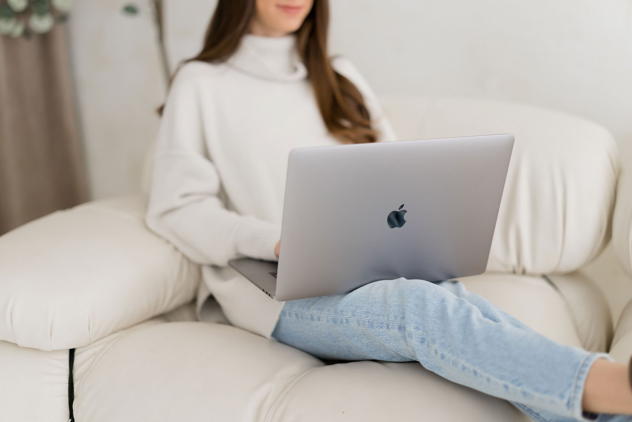A person sitting on a cream-colored sofa using a silver MacBook laptop, wearing a white sweater and light blue jeans.
