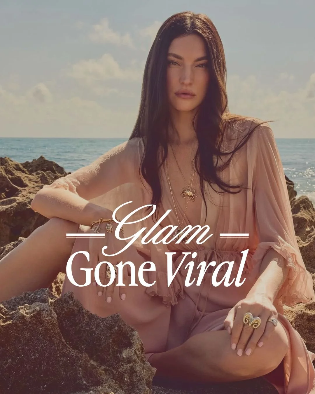 A woman sitting on rocks at the beach wearing a pink dress, with jewelry including rings, necklaces, and a bracelet, with the sea and sky in the background.
