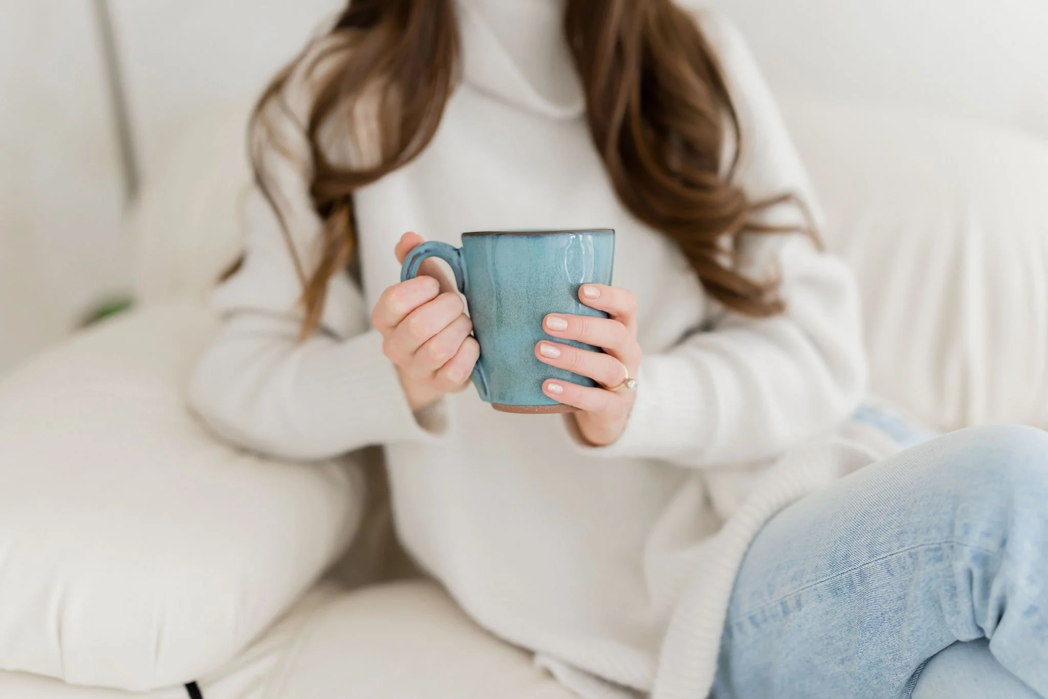 A woman in a white sweater sitting on a cream-colored sofa holding a blue ceramic mug.