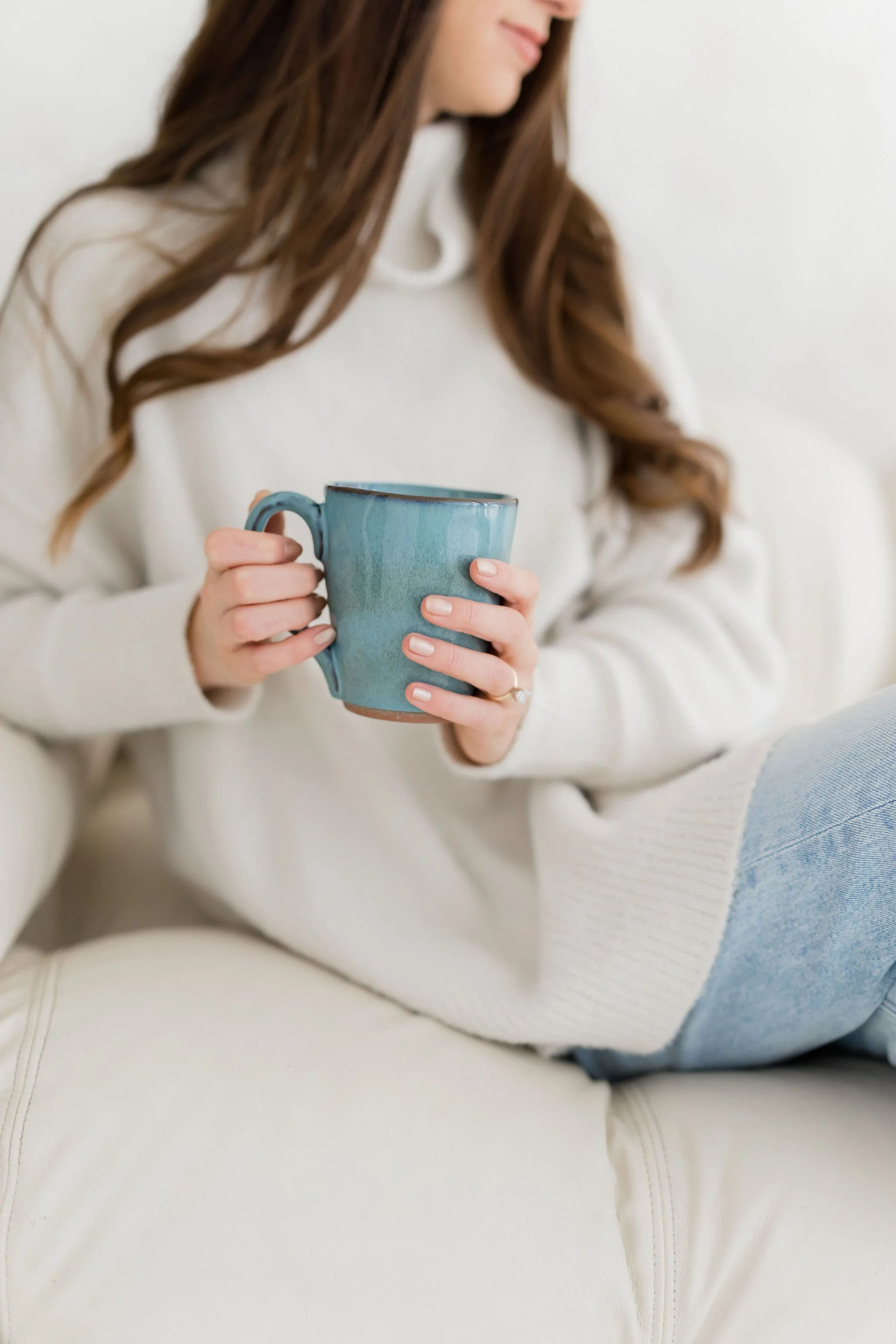 A woman with long brown hair wearing a white sweater and light blue jeans, sitting on a cream-colored couch, holding a turquoise mug with both hands.