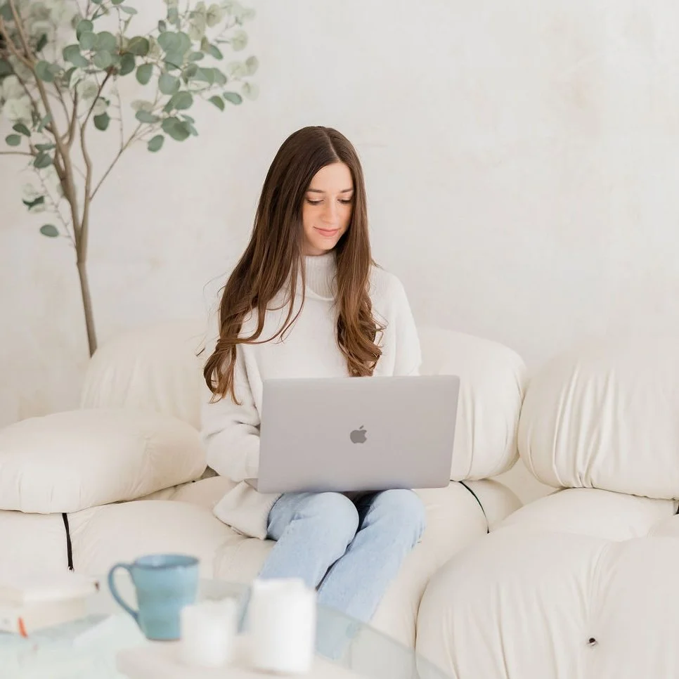 A young woman with long brown hair sitting on a white couch using a silver MacBook laptop, with tea cups and a plant in the background.
