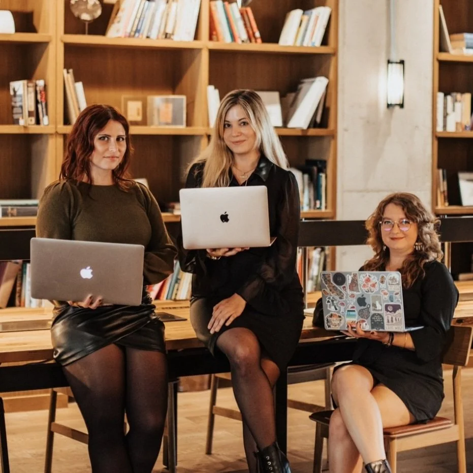 Three women sitting and standing in a library or bookstore with wooden shelves filled with books behind them, each holding a laptop.