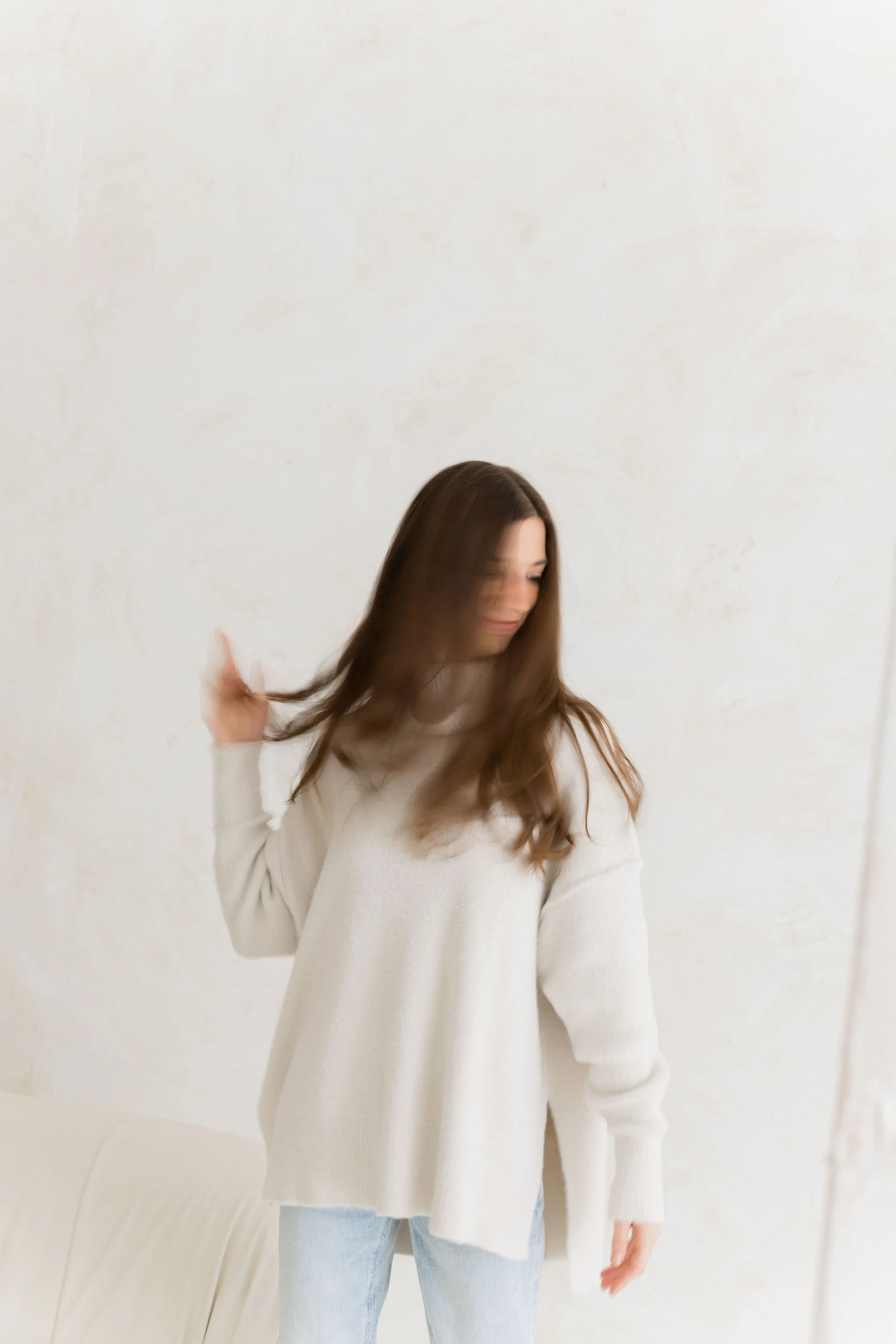 Woman with long brown hair wearing a cream-colored sweater and light blue jeans, standing against a plain white wall, brushing her hair.
