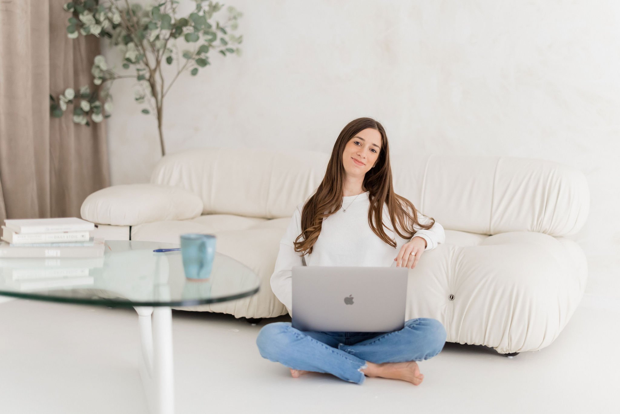 A woman with long brown hair sits cross-legged on a white carpeted floor, working on a silver MacBook laptop. She is wearing a white sweater and blue jeans, and he has a slight smile. The setting is a bright, minimalist living room with a white sofa, a glass coffee table, some books, a blue mug, and a potted plant near the wall.