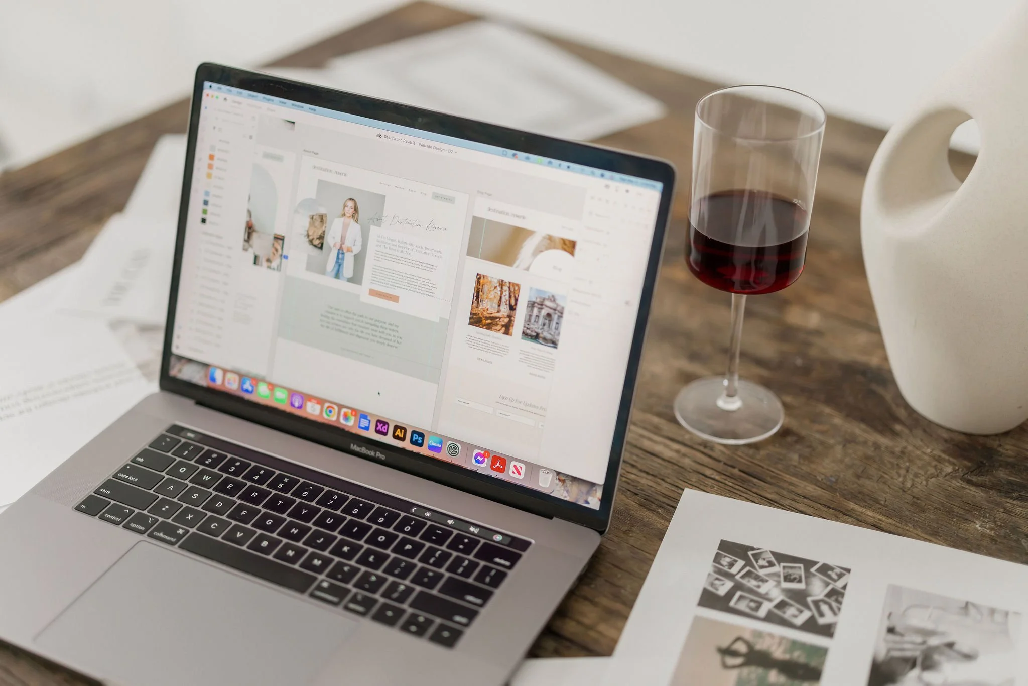 A MacBook Pro laptop on a wooden table displaying a website design project with tools and images. Next to the laptop is a glass of red wine and a white ceramic vase. Papers with black-and-white photographs are also on the table.