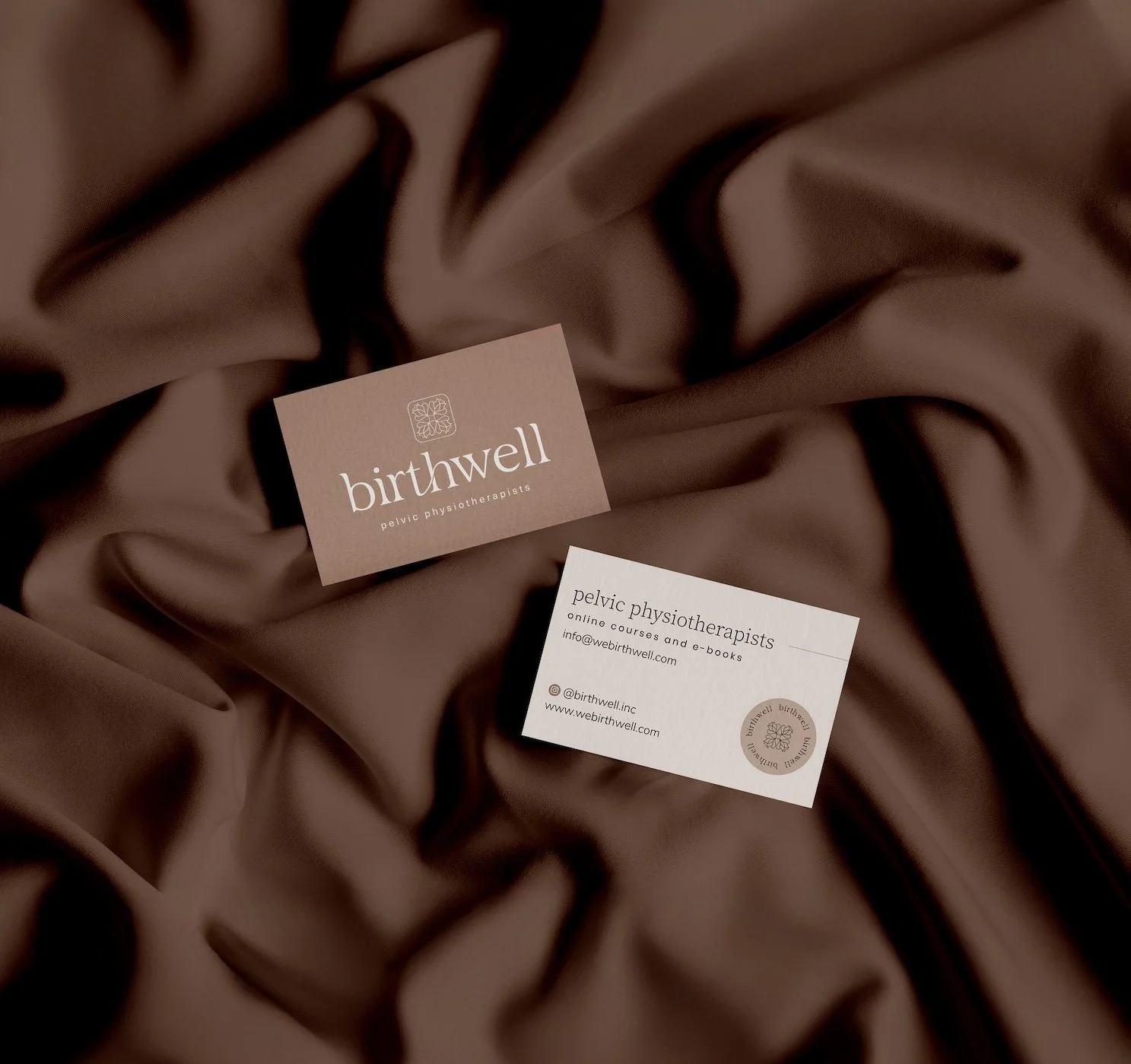 Business cards for Birthwell, a pelvic physiotherapy clinic, placed on a brown satin fabric background.