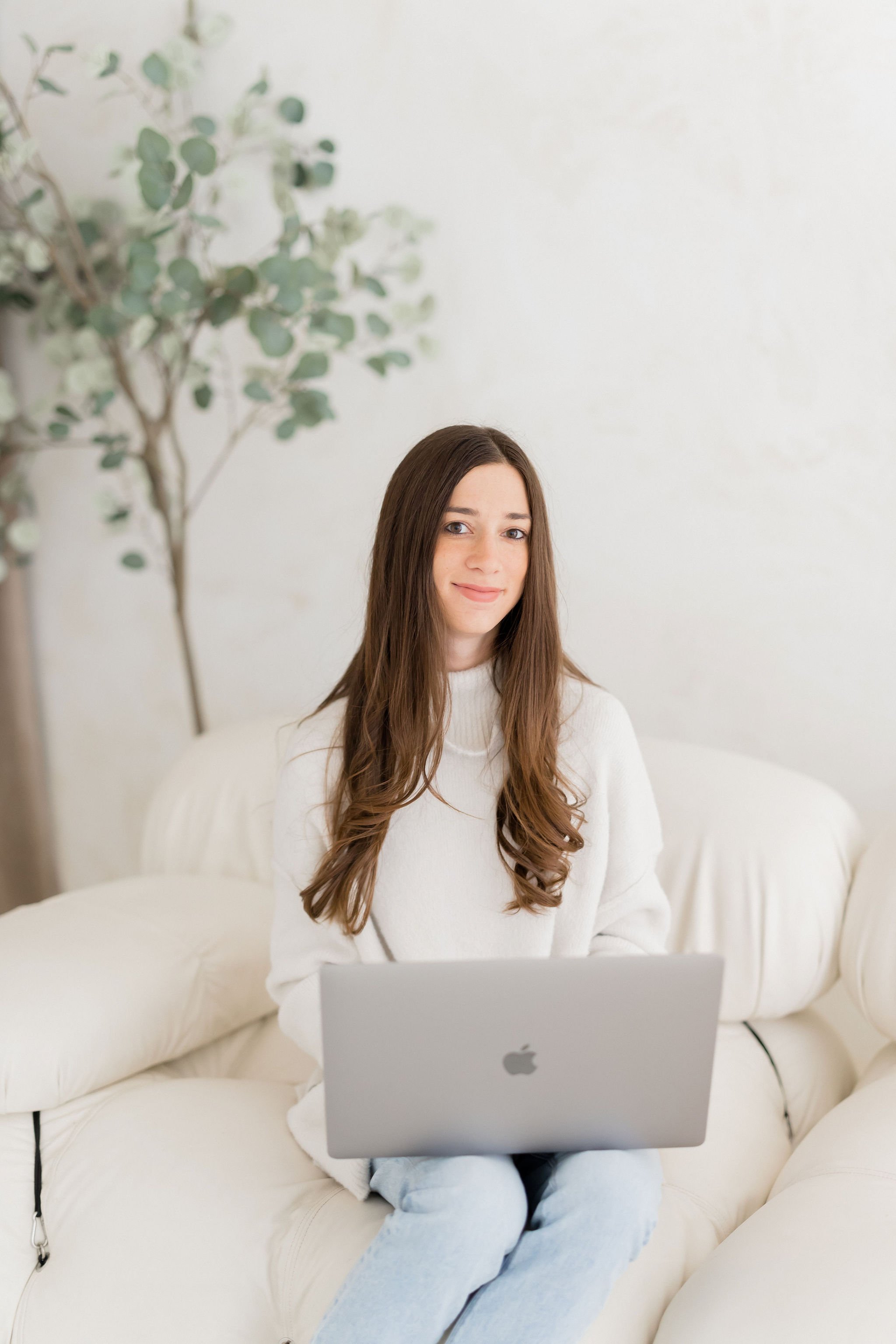 A young woman with long brown hair using a silver MacBook while sitting on a white sofa with a plant in the background.