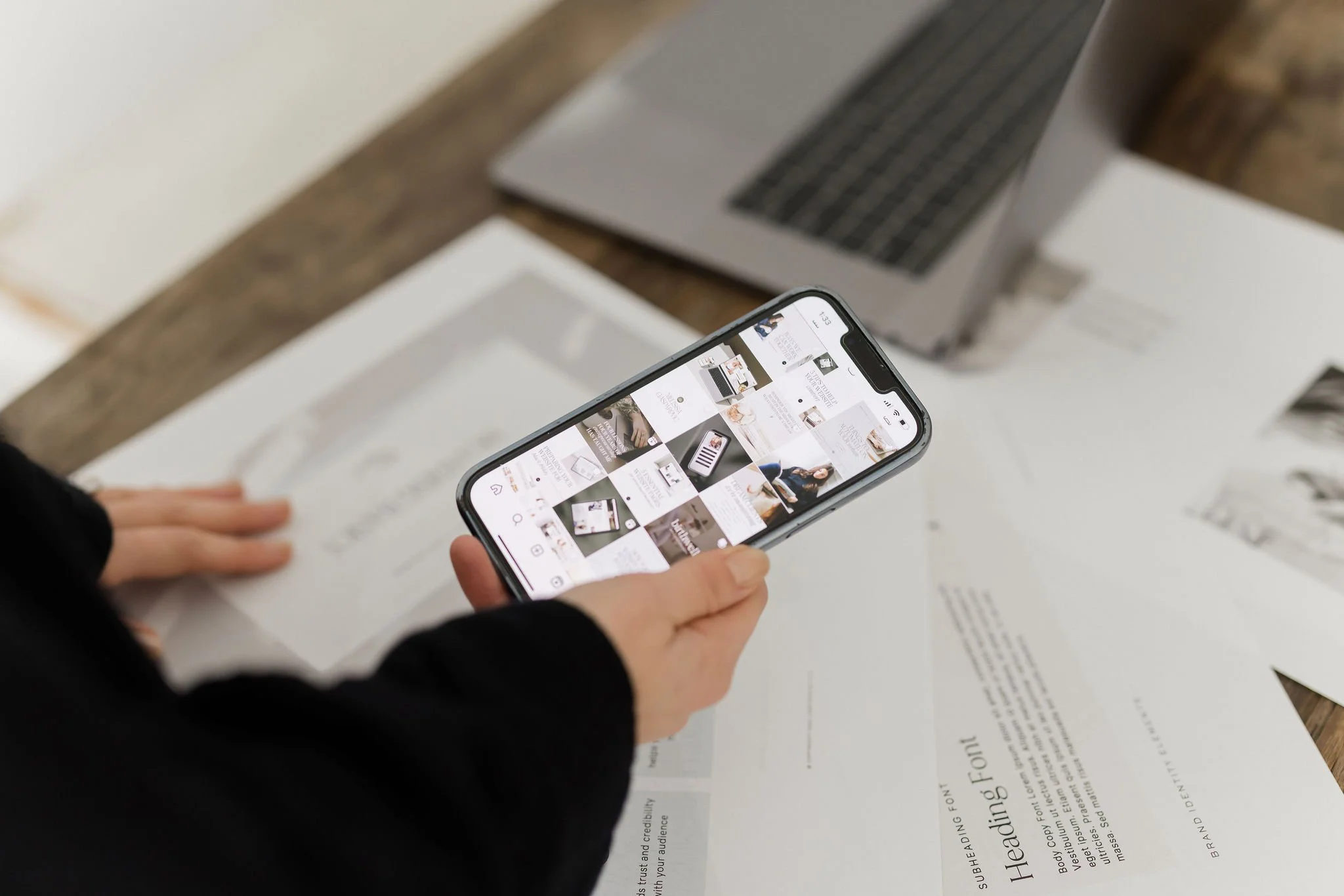 Person holding a smartphone, viewing an Instagram grid, on a wooden table with papers and a laptop.