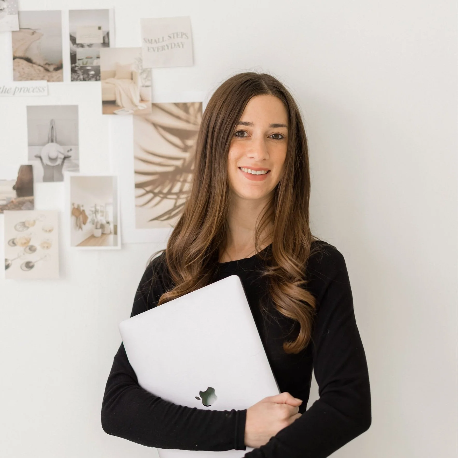 A woman with long brown hair holding a silver MacBook in front of a white wall with framed photos and quotes.