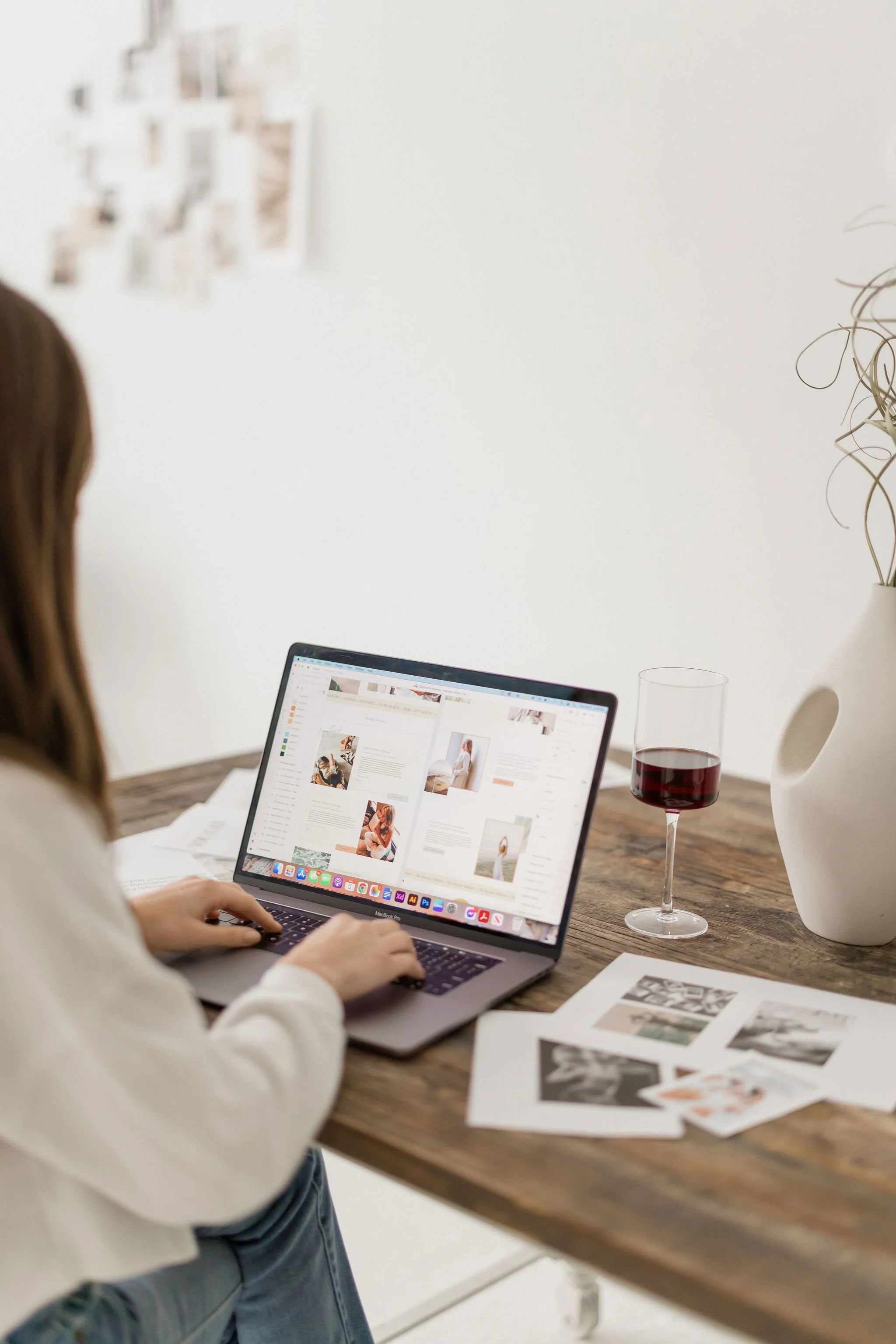 A woman working on a laptop at a wooden desk, with printed photographs and a glass of red wine nearby, in a bright room.