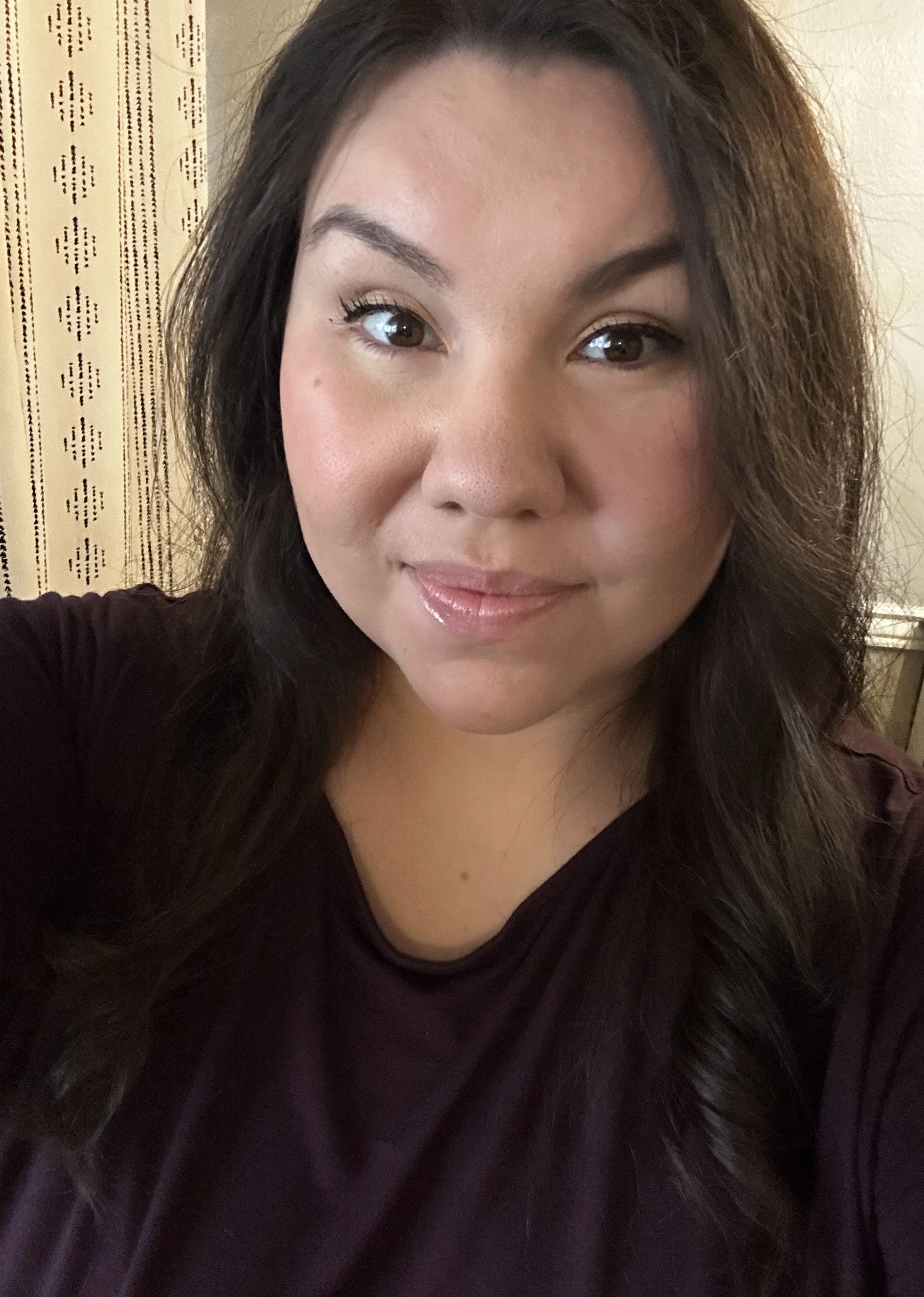 A woman with long dark hair and makeup taking a selfie indoors, wearing a dark top.