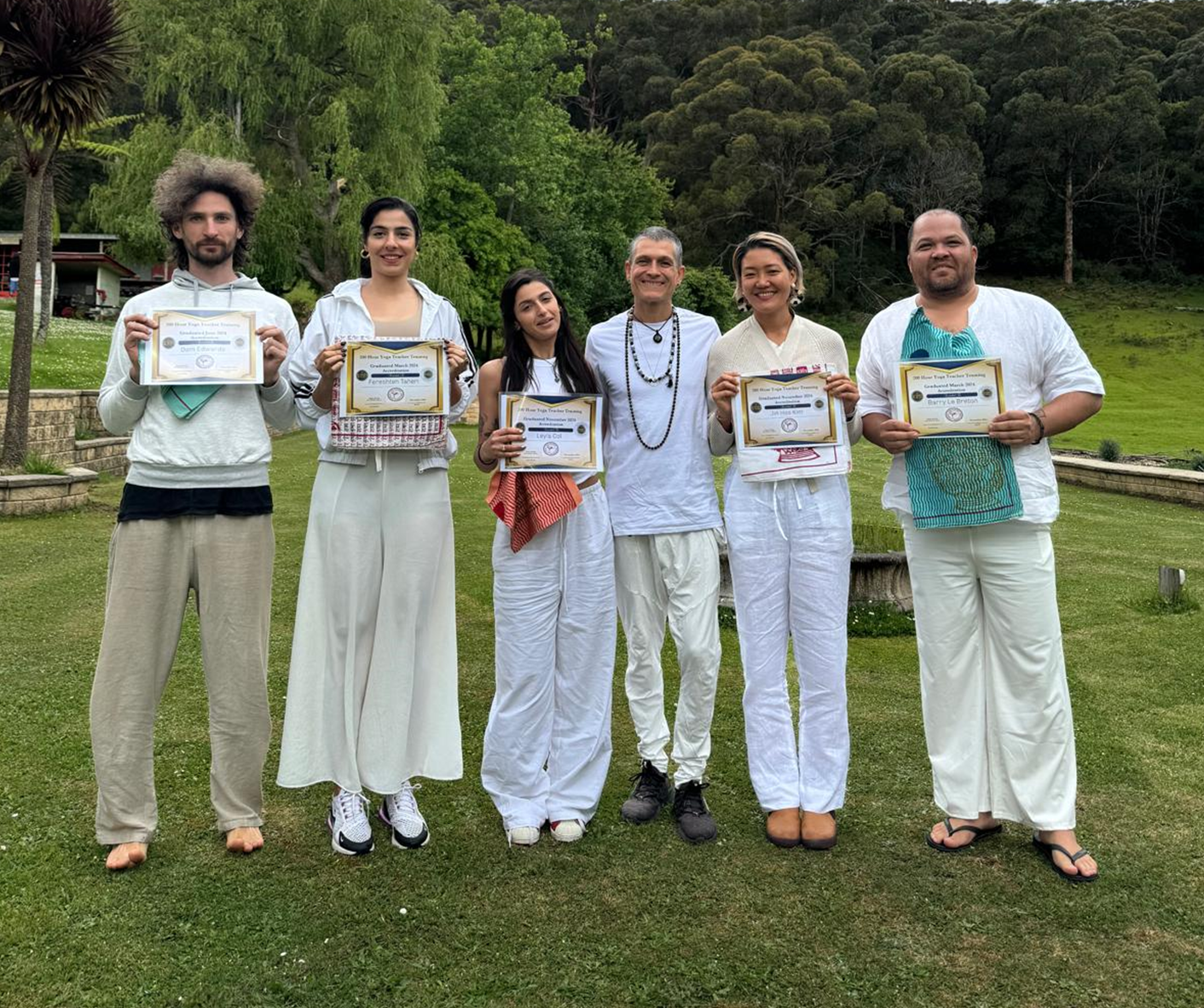 Group of people outdoors in white clothing holding certificates, standing on grass with trees in the background.