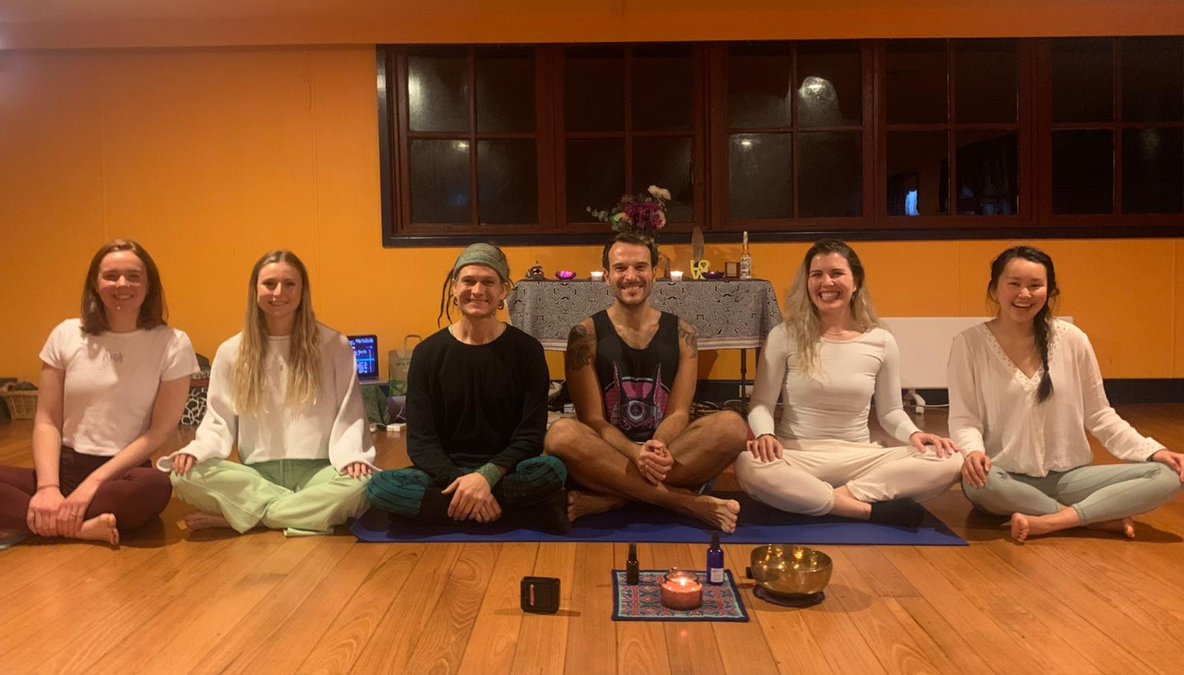 Group of six people sitting cross-legged on a wooden floor with a small table in the background, candles and a singing bowl in the foreground.
