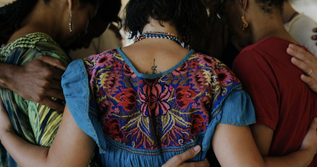 A group of people embracing, featuring a woman in a colorful embroidered blouse.