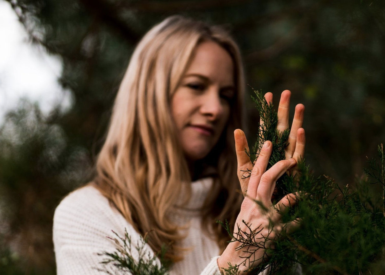 Woman in white sweater touching pine branches outdoors.