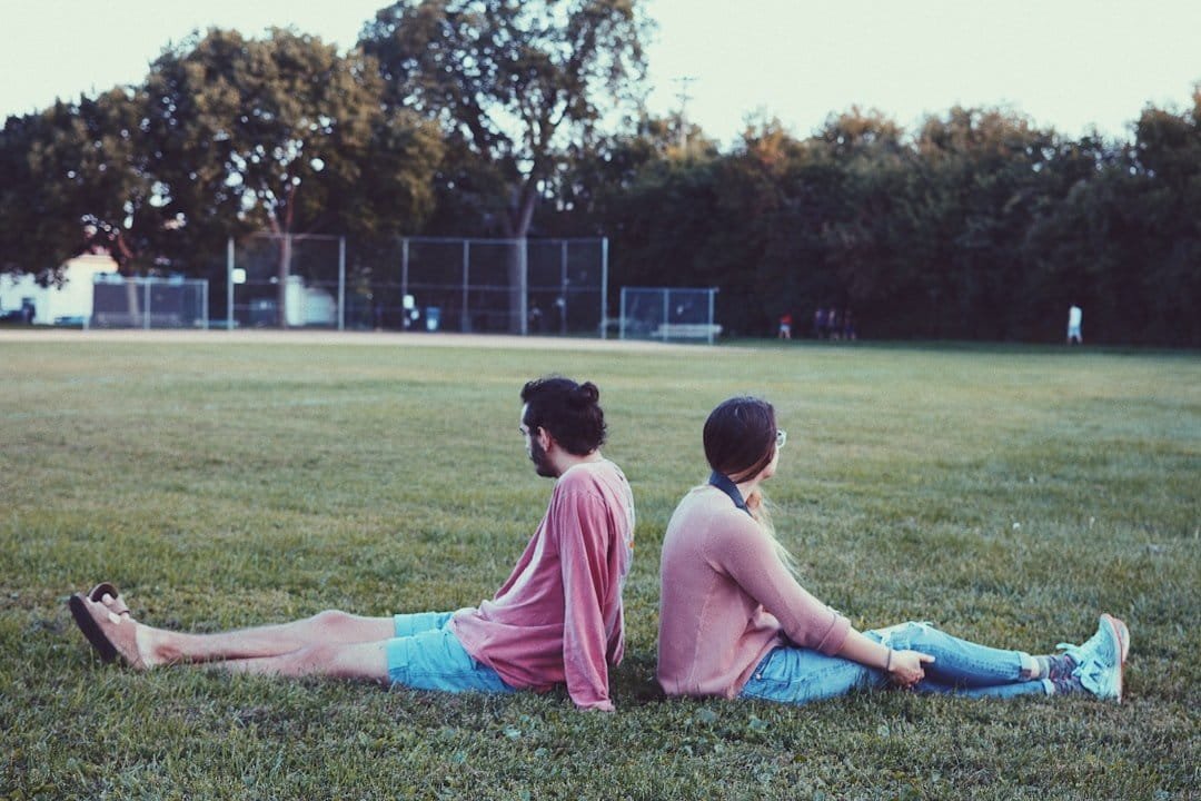 Man and a woman sitting on grass facing away from each other wearing pink shirts and blue jeans.