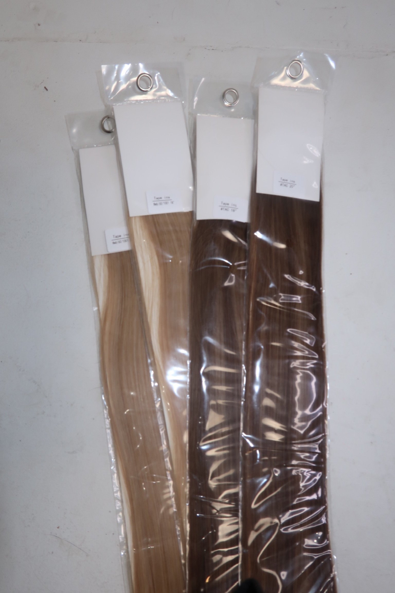 Four hair extensions in varying shades of blonde and brown, sealed in plastic packaging, hanging against a white wall.