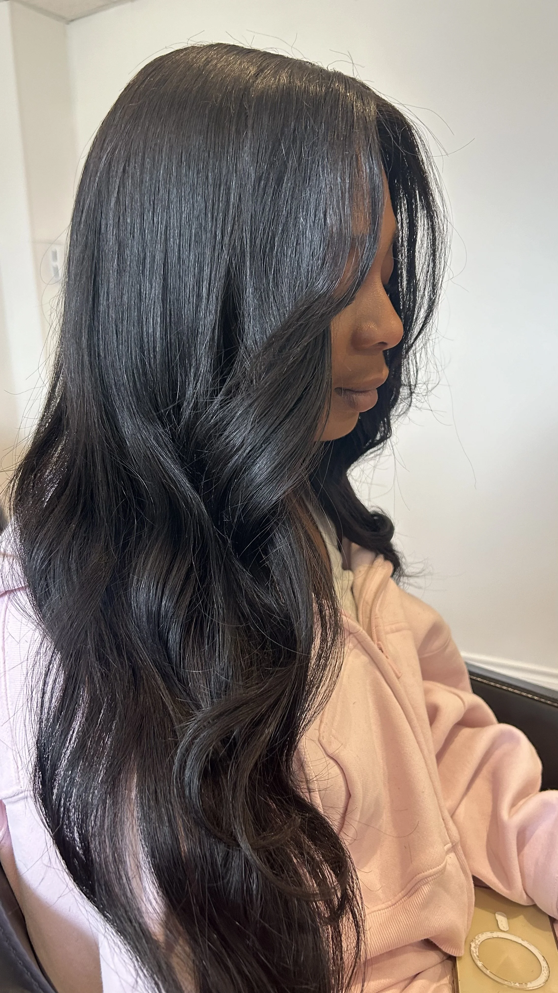 Side profile of a woman with long, wavy black hair, sitting indoors near a plain white wall, wearing a light pink hoodie, with a bracelet on her wrist and a smartphone on her lap.