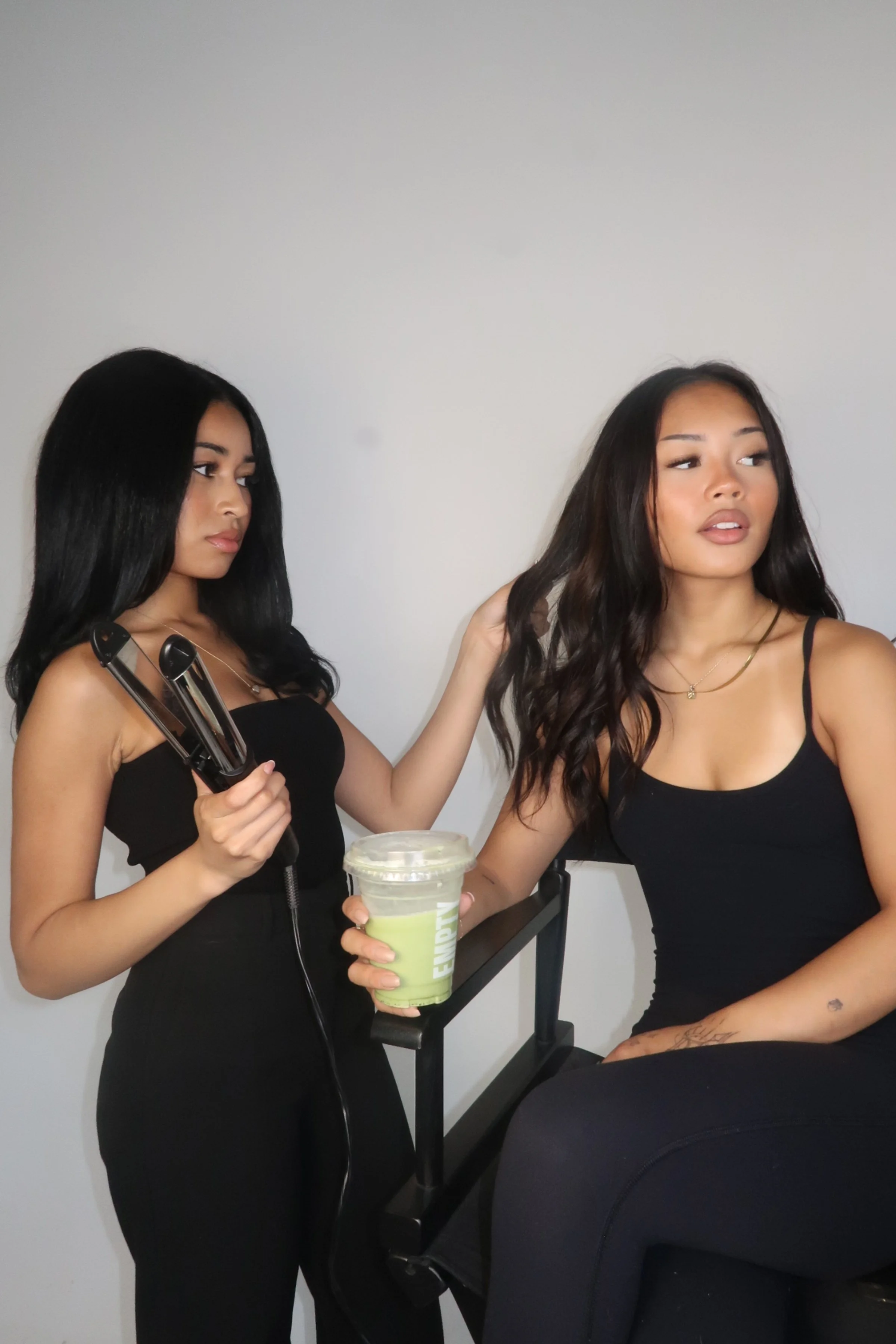 Two women in black tops, one with long black hair holding a curling iron, the other with long wavy hair sitting on a salon chair, holding a green beverage, with a plain white wall in the background.