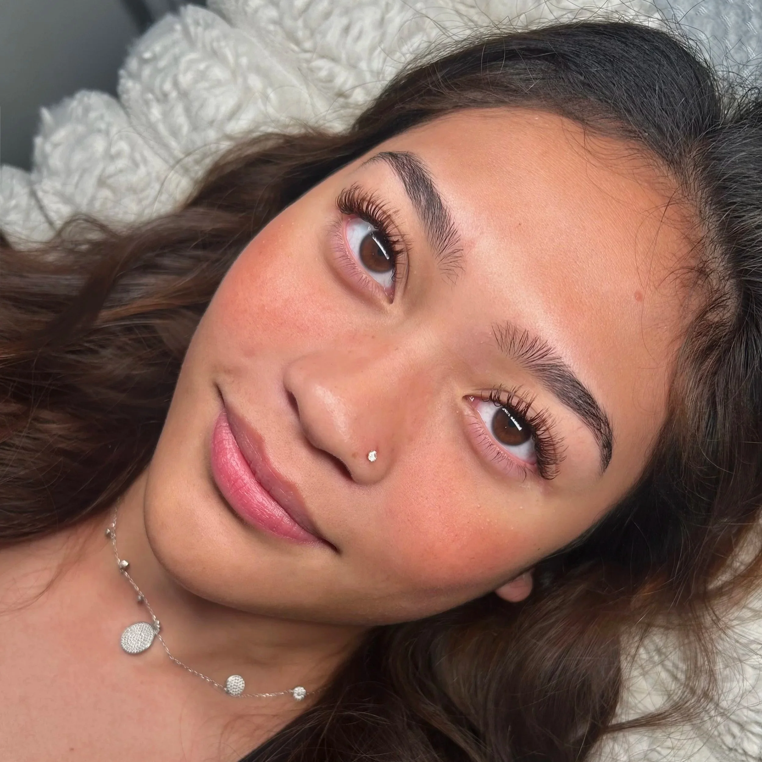 Close-up of a young woman with brown hair and light brown eyes, lying on a white fluffy pillow, smiling softly, wearing a silver necklace and a small nose stud.