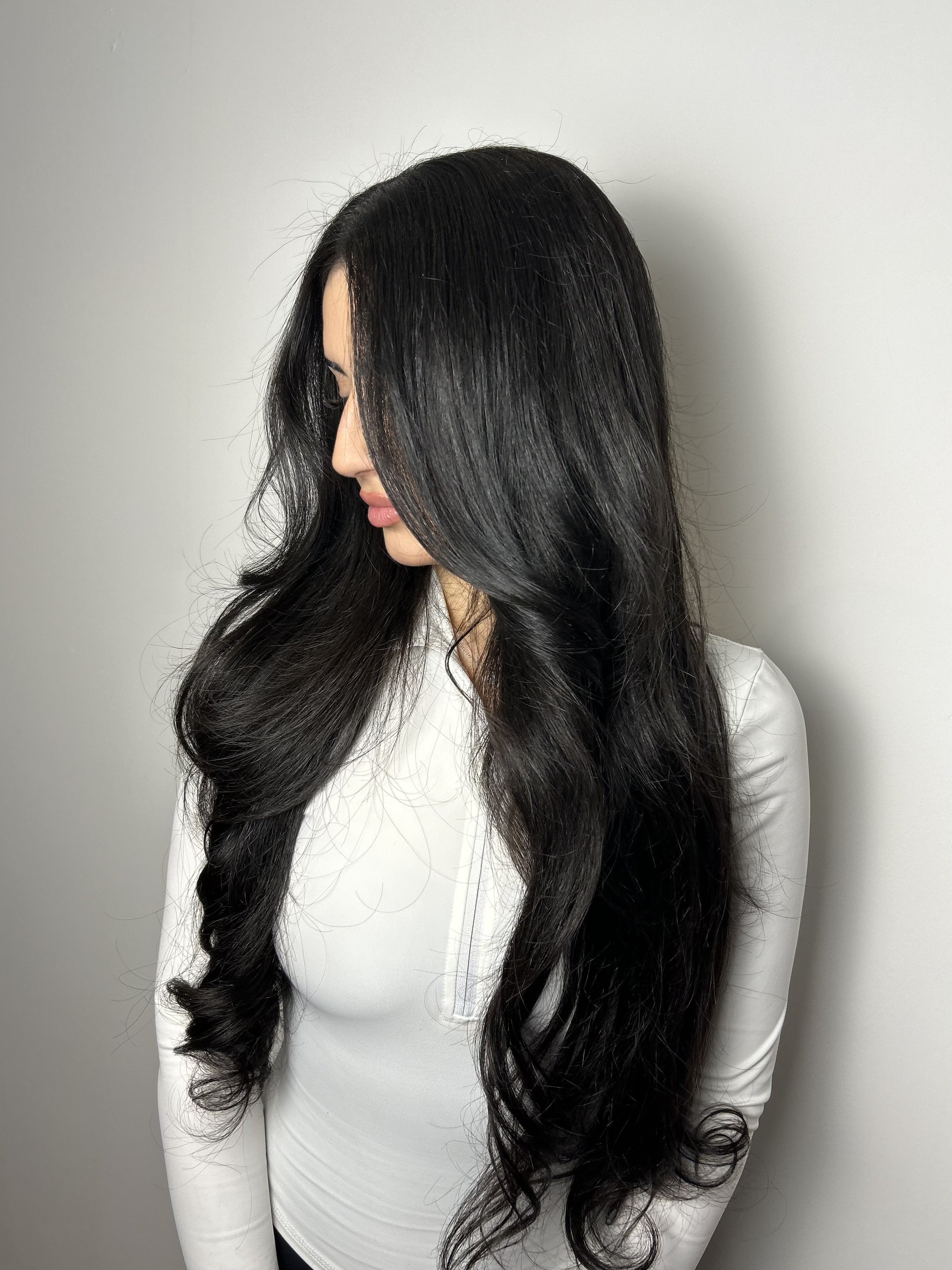 A woman with long, wavy black hair, wearing a white top, is standing against a plain light gray background, with her head turned to the side.