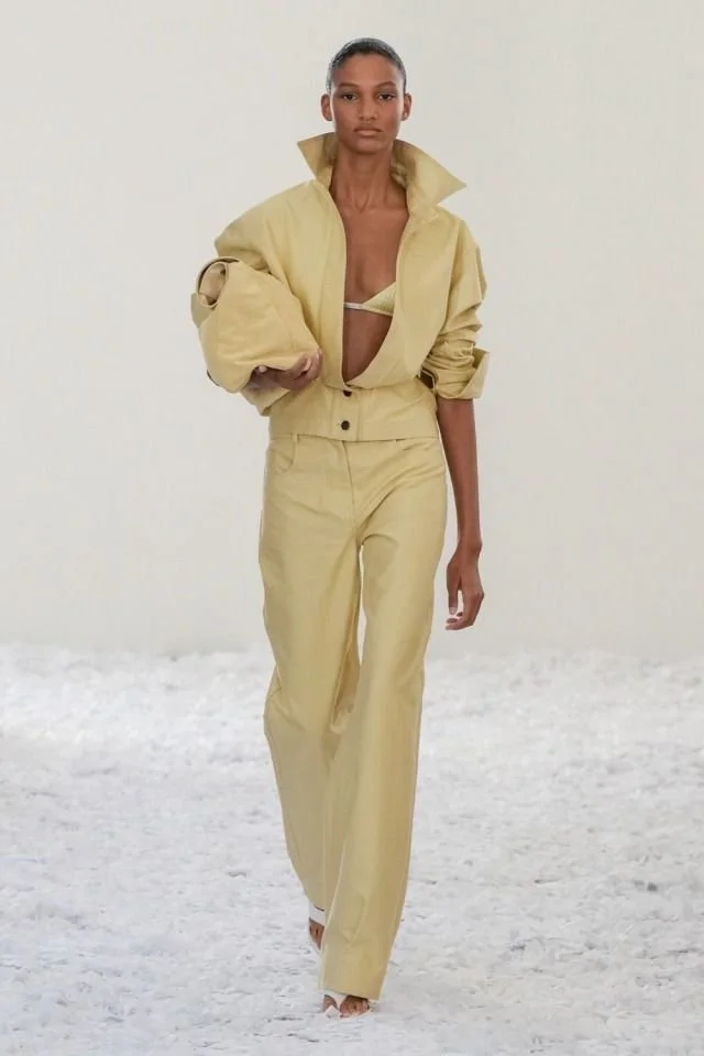 A fashion model walking on a runway wearing a beige outfit consisting of high-waisted pants and a matching oversized jacket, with a beige top underneath, against a neutral background.