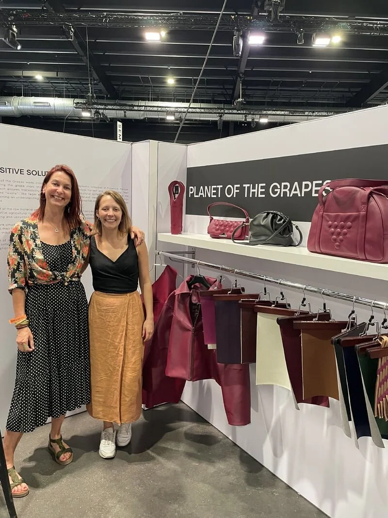 Two women standing in front of a display of handbags and fabric samples at the 'Planet of the Grapes' booth at an event or exhibition.