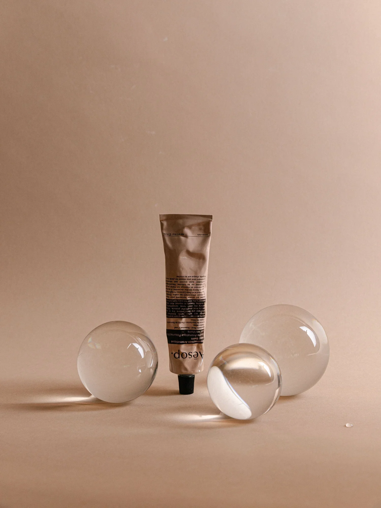 A tube of beige cream or paint surrounded by four glass marbles on a beige surface with a plain beige background.