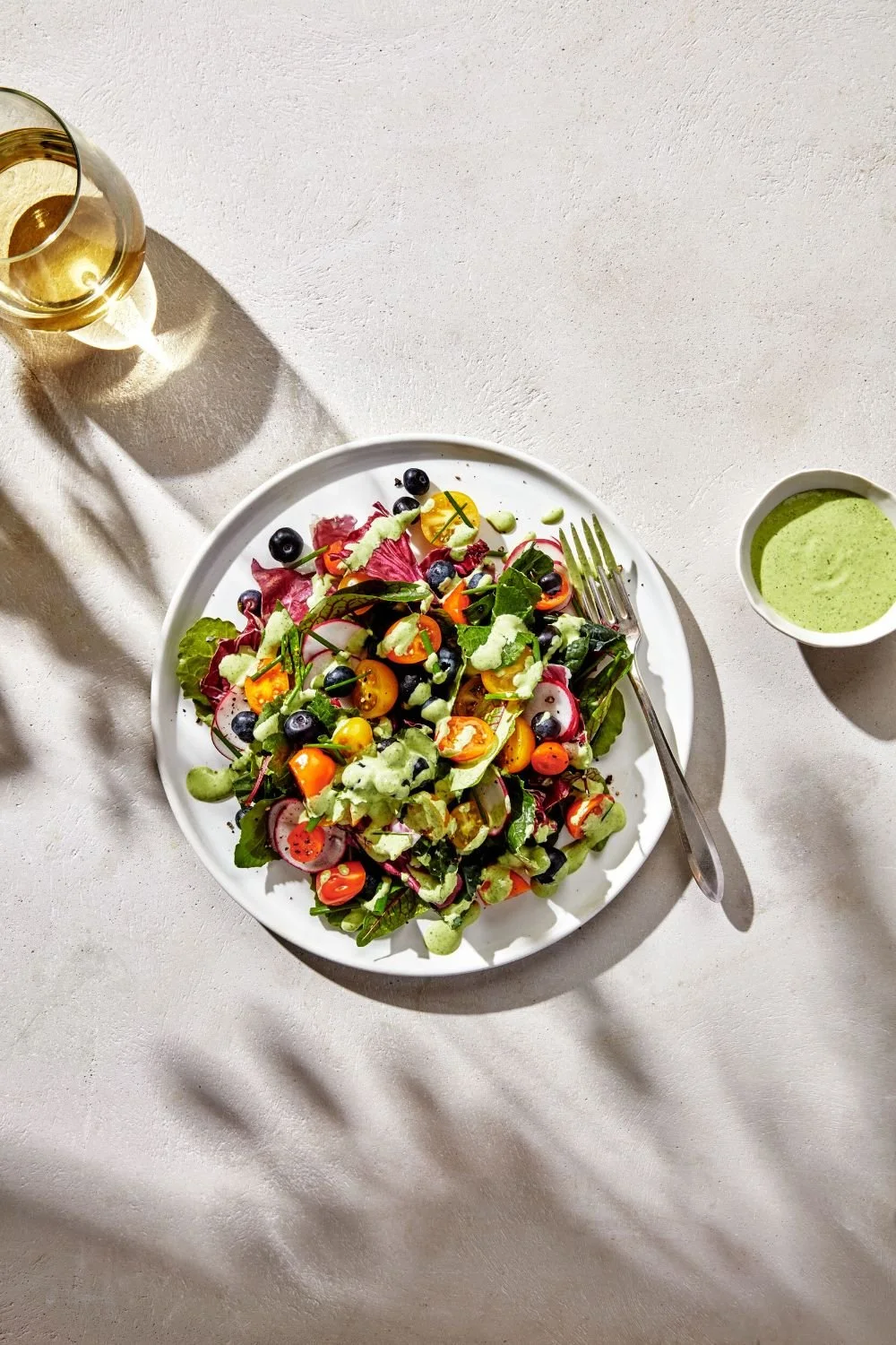 A plate of mixed salad with cherry tomatoes, radishes, and blueberries, served with a side of green salad dressing, a glass of white wine, and a small bowl of green dressing on a white table.