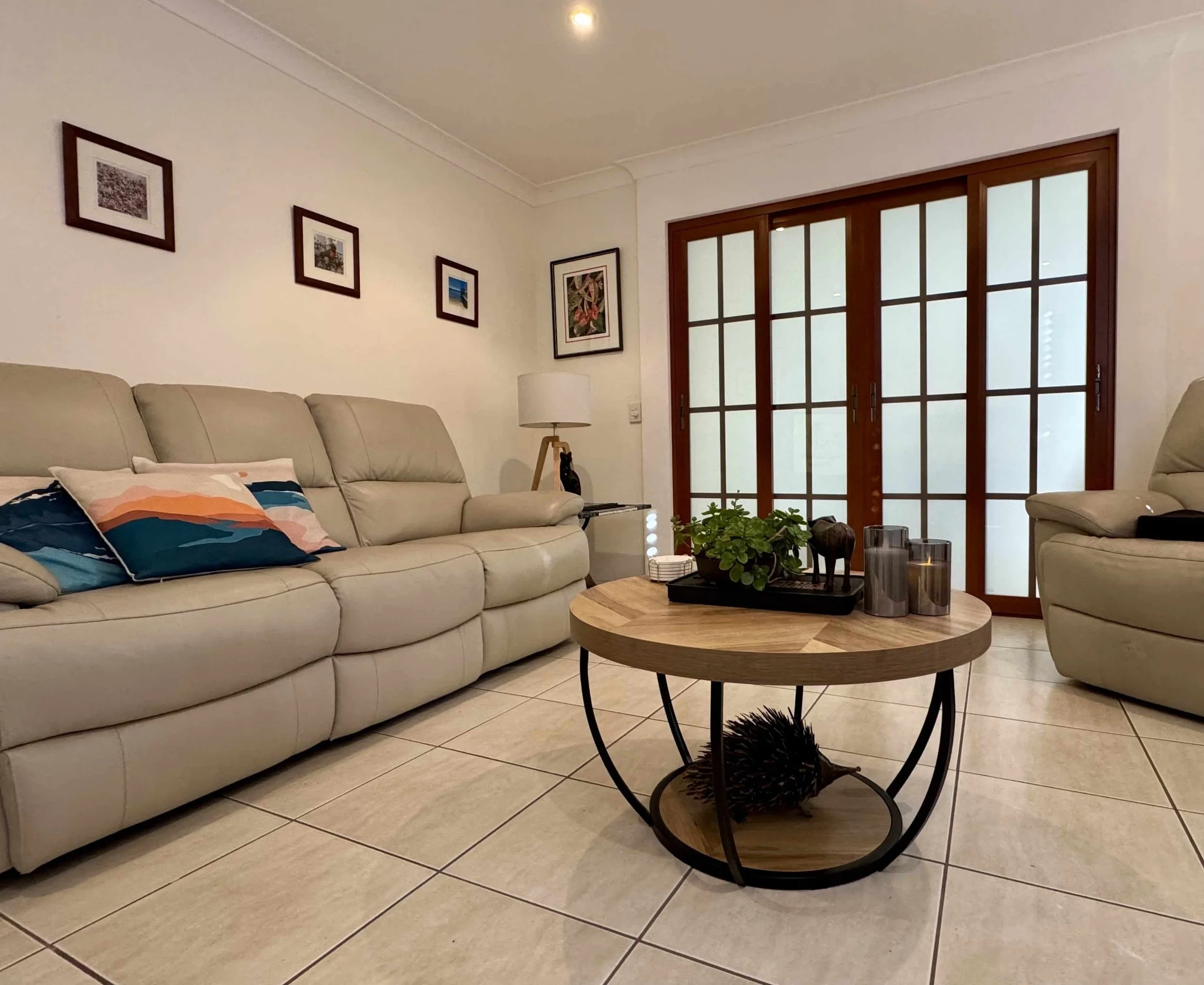 Living room with beige leather sofa, armchair, round wooden coffee table with plants and decorative items, tiled floor, white walls with framed pictures, and sliding glass doors with wooden frames.
