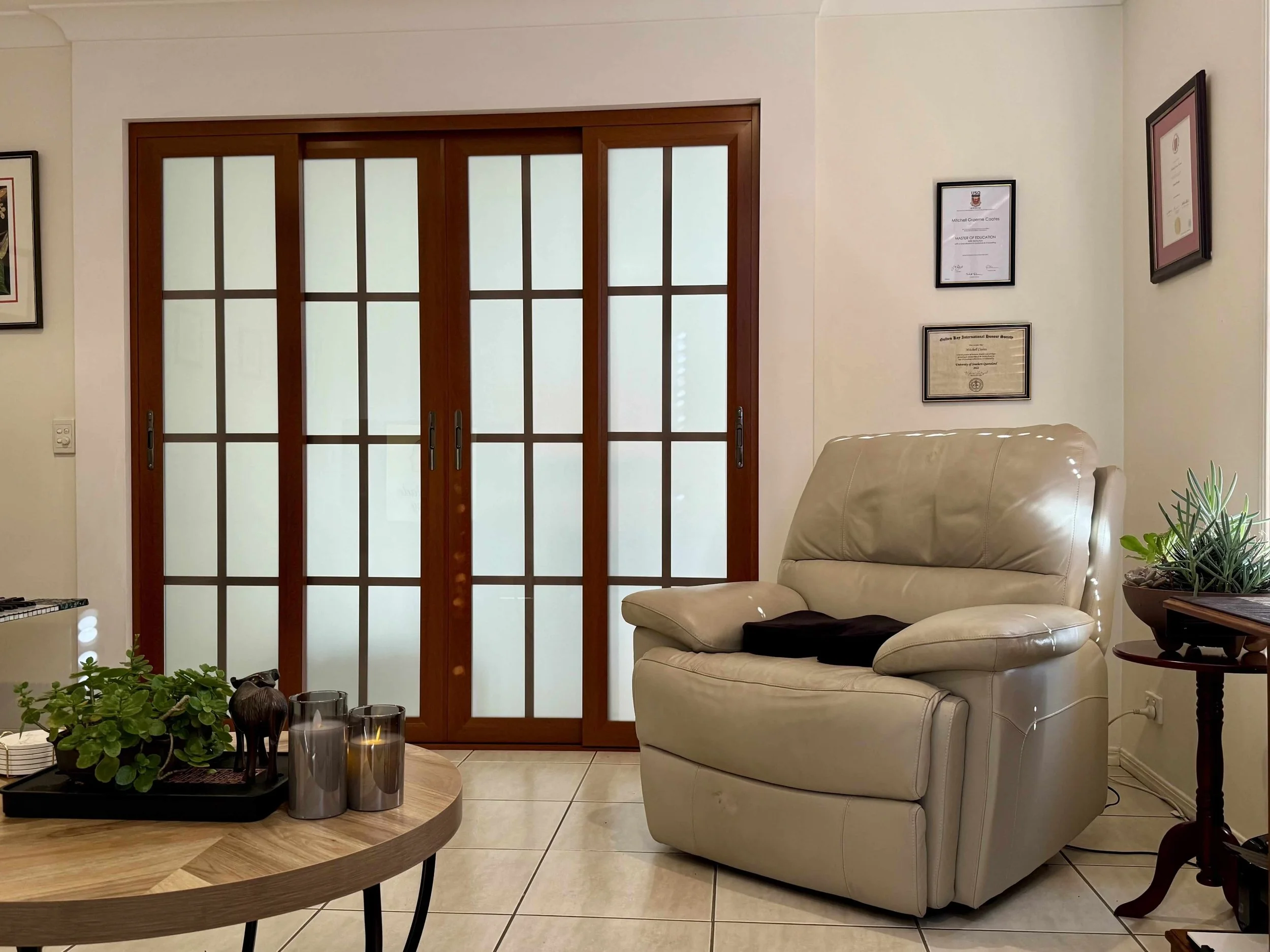 Living room with beige leather recliner, wooden coffee table with plants and candles, frosted glass sliding doors, framed certificates and artwork on wall, potted plants on side tables.