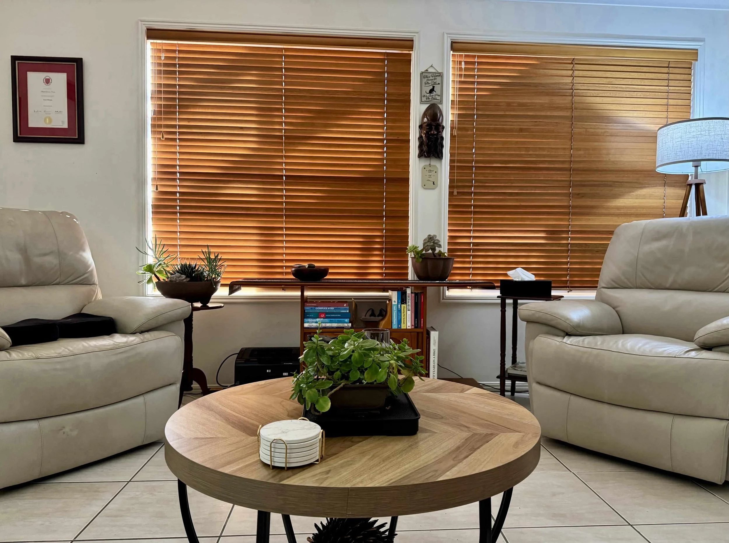 Living room with two beige leather sofas, wooden blinds on windows, wooden coffee table with a plant, and various decorative items and books.