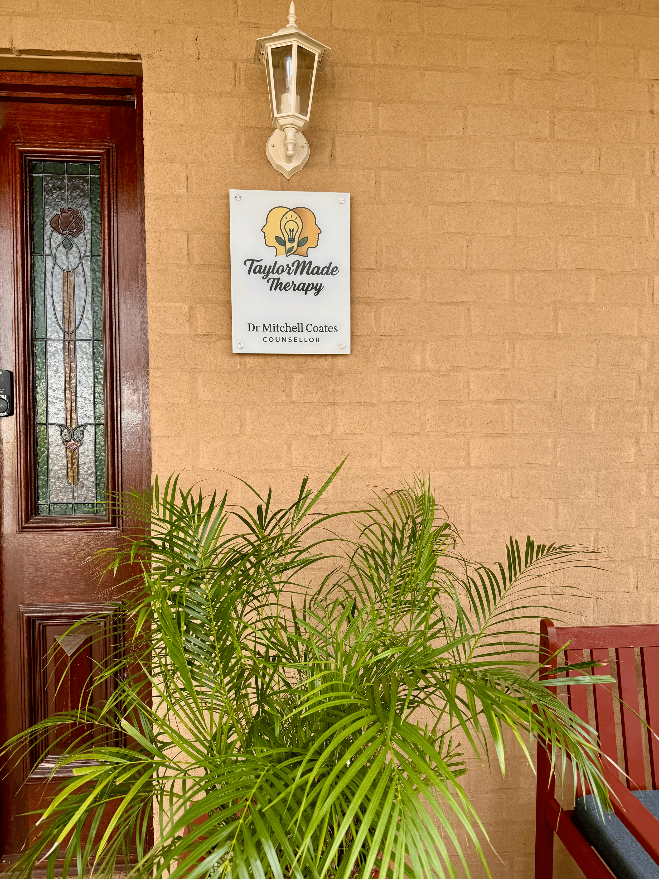 Sign for TaylorMade Therapy and Dr. Mitchell Coates, counsellor, mounted on brick wall beside door with stained glass, bench, and potted plant.