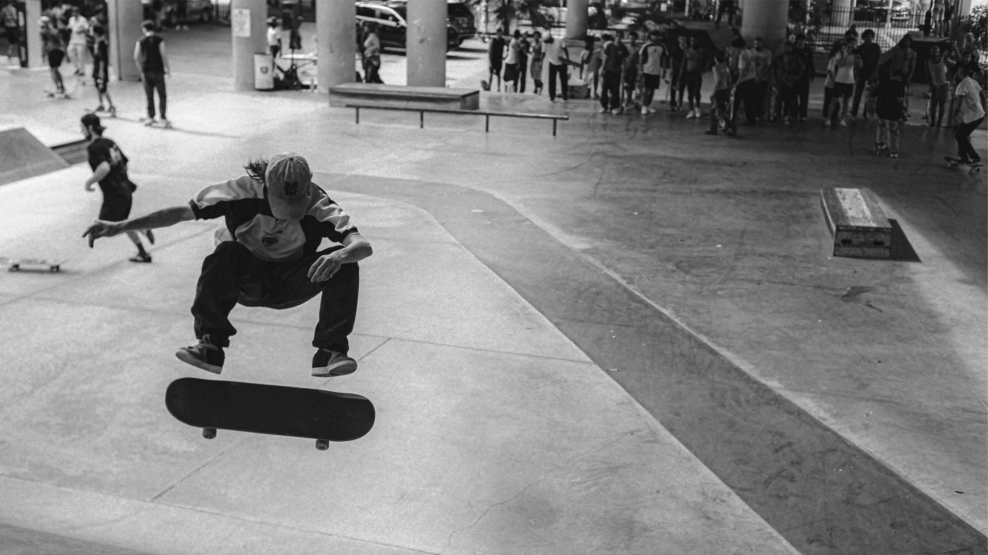 A skateboarder performing a trick at a skate park, with onlookers and other skaters in the background.