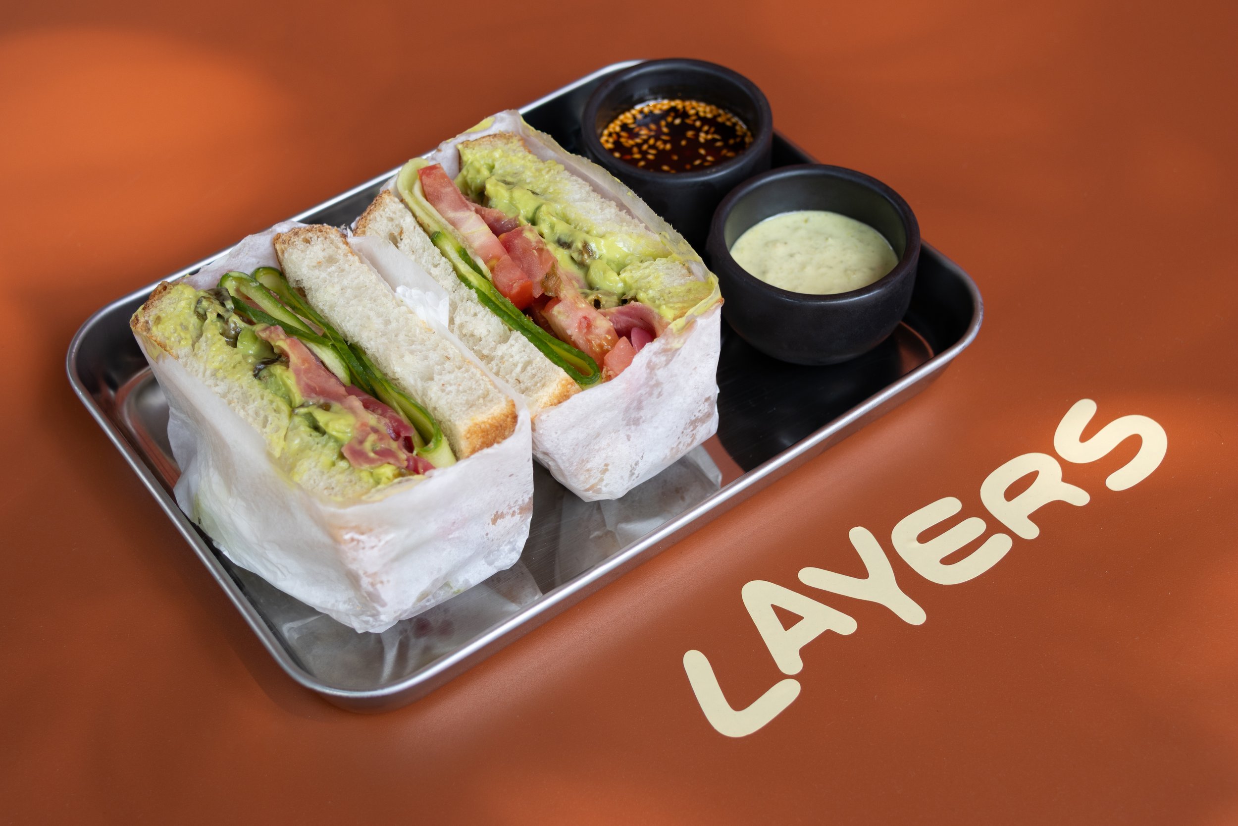 Sándwich de pan blanco con aguacate, tomate, cebolla y jamón, acompañado de dos salsas en pequeños tazones en una bandeja de metal sobre una mesa anaranjada con la palabra "LANERS".