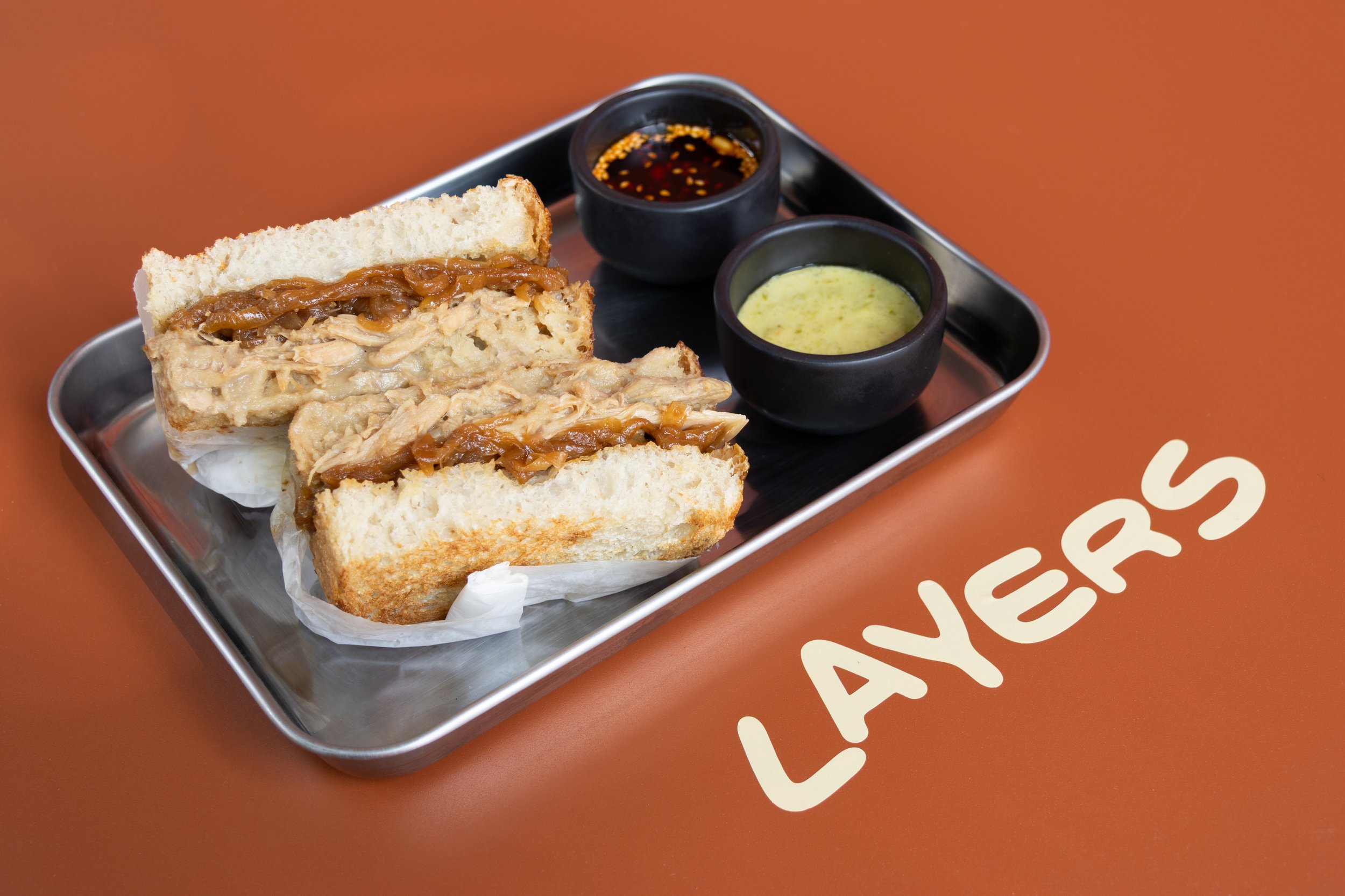 Sandwich de pollo con cebolla caramelizada en plato de metal, acompañado de salsa de soya y salsa de jengibre en pequeños tazones negros, sobre mesa naranja con la palabra 'LA YERAS'.