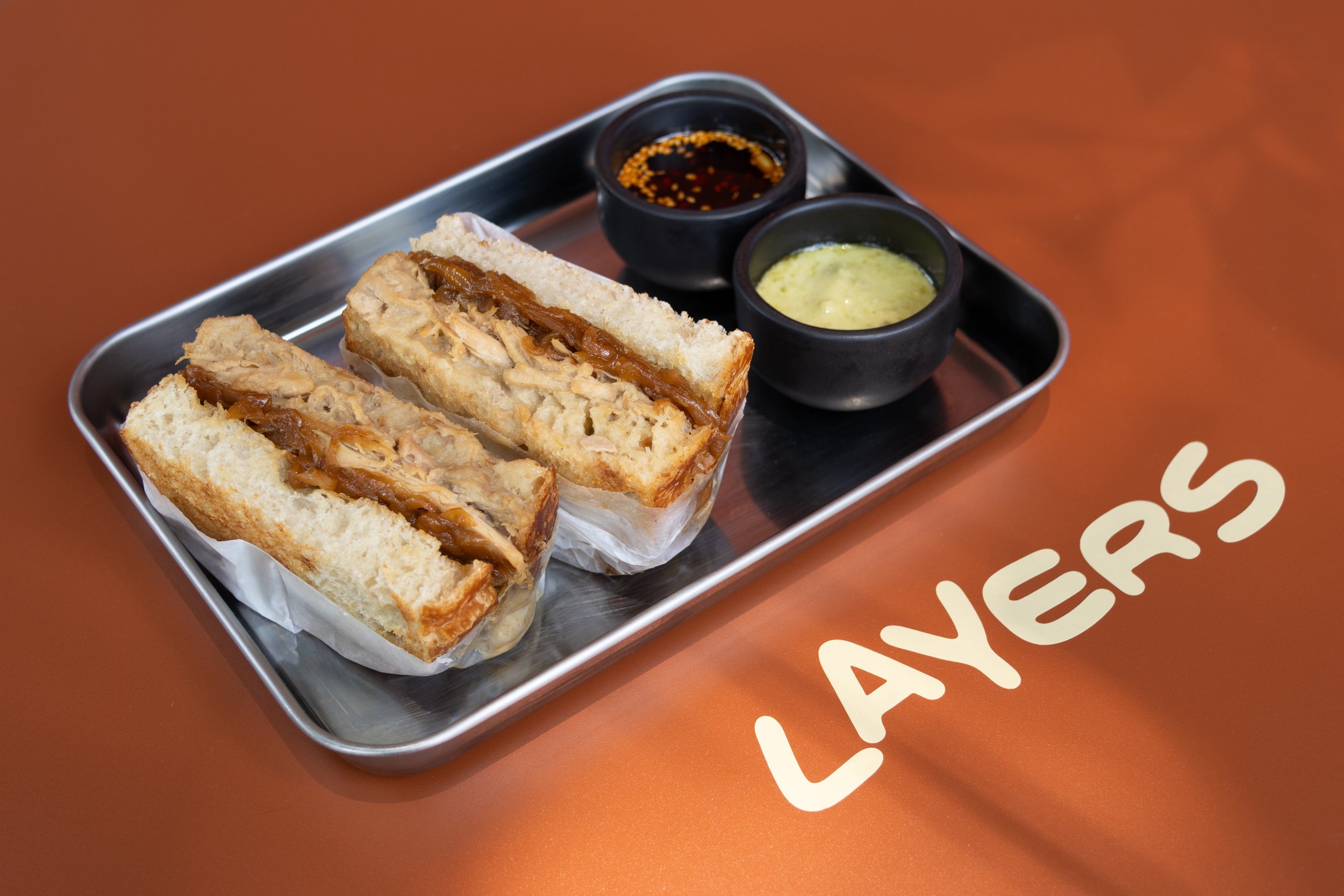Sandwich de pollo con cebolla y salsas en plato de metal, sobre mesa color naranja con la palabra "LAYERS".