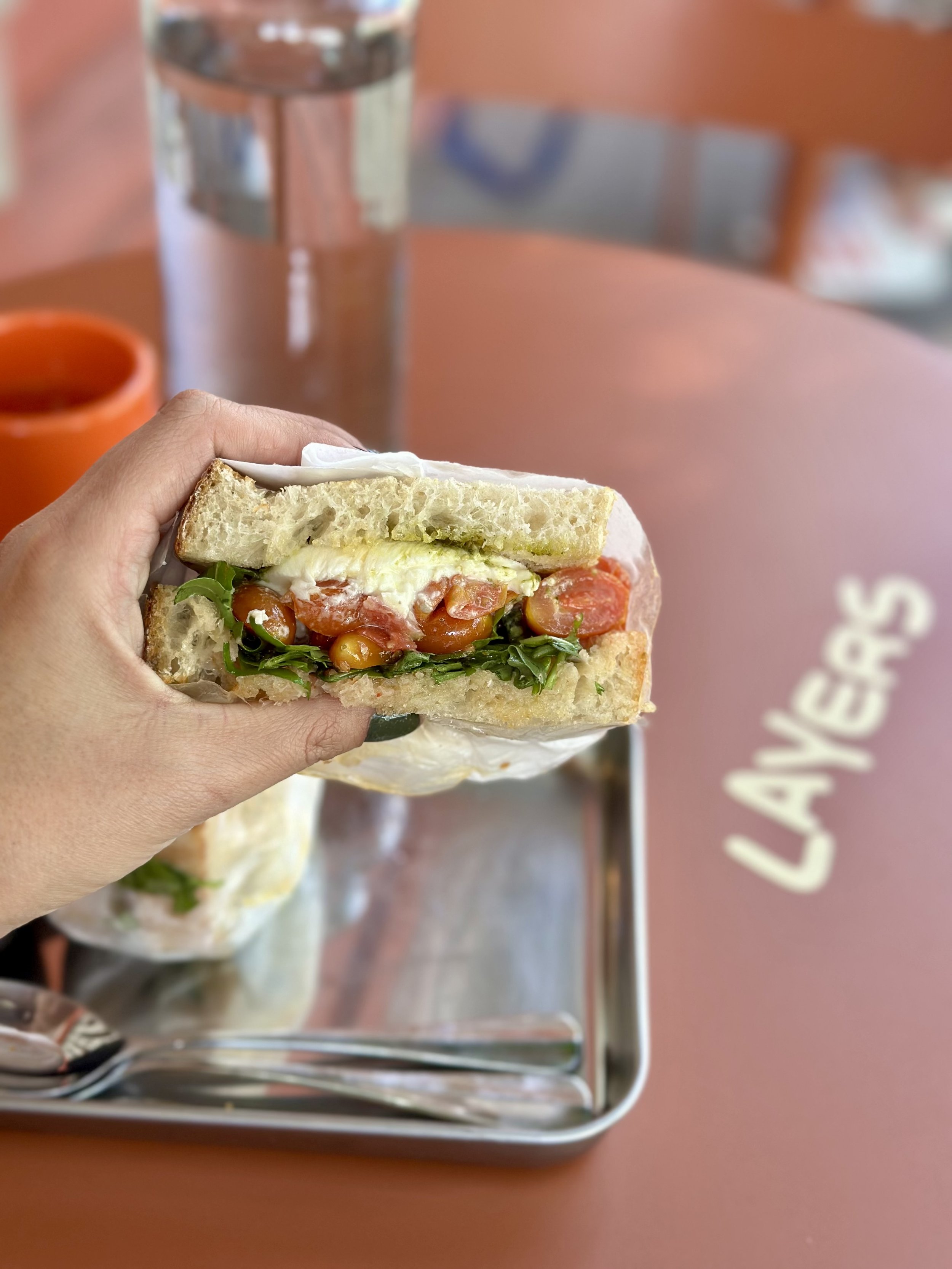 Un sándwich de pan blanco con tomate, lechuga, mayonesa y otros ingredientes, sostenido por una mano. En el fondo, una taza naranja, un vaso de agua y una mesa de color terracota con la palabra 'LAVERS' escrita.