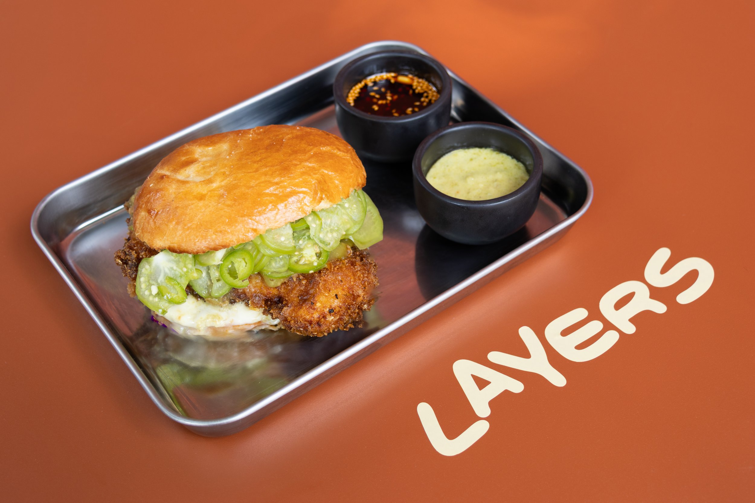 Sandwich con pollo empanizado, cebollín, y salsas en plato de aluminio, con la palabra 'LAVESAS' escrita en la mesa