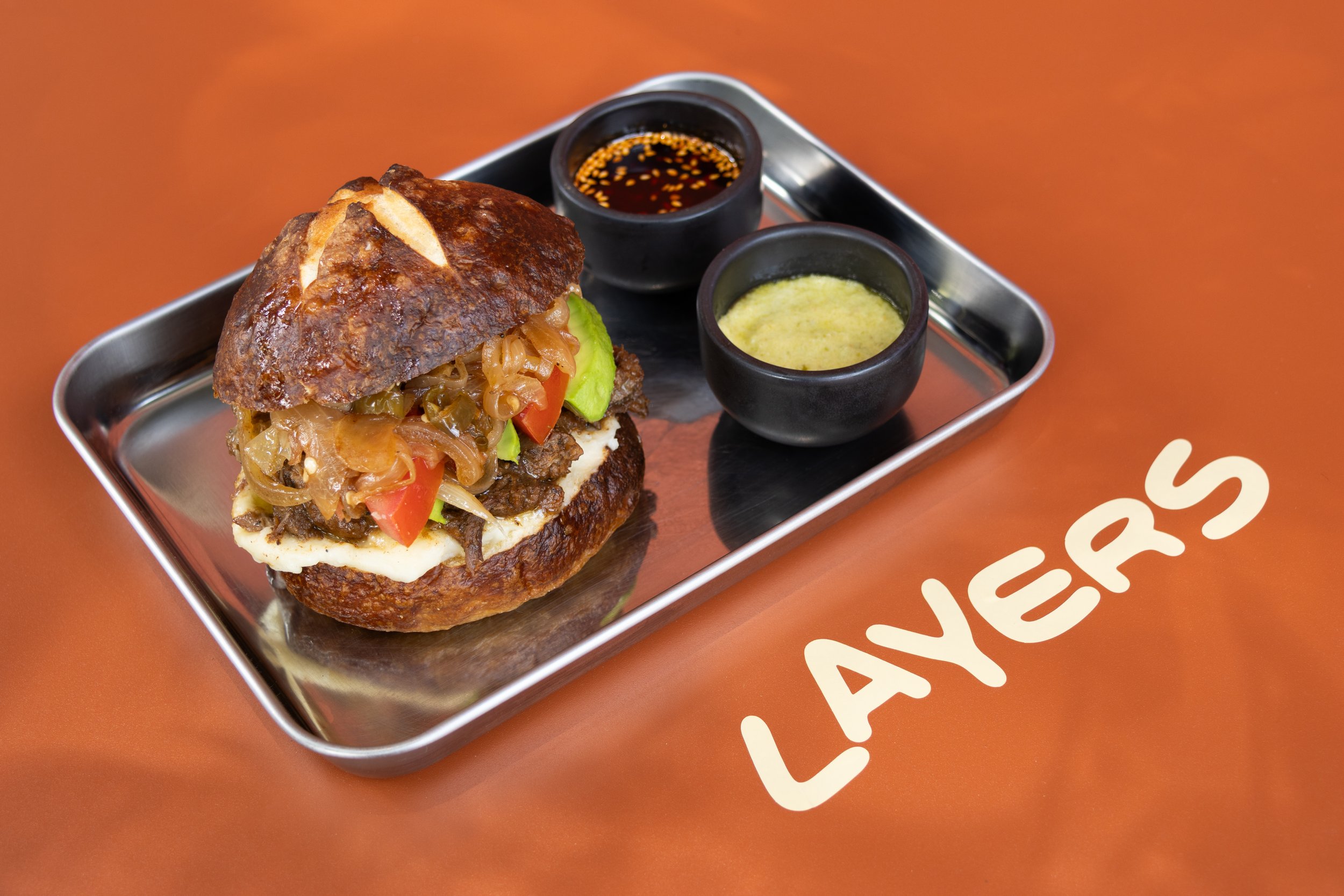 Sandwich de carne con verduras y salsas en bandeja de metal sobre mesa con fondo naranja y la palabra LAYERS.