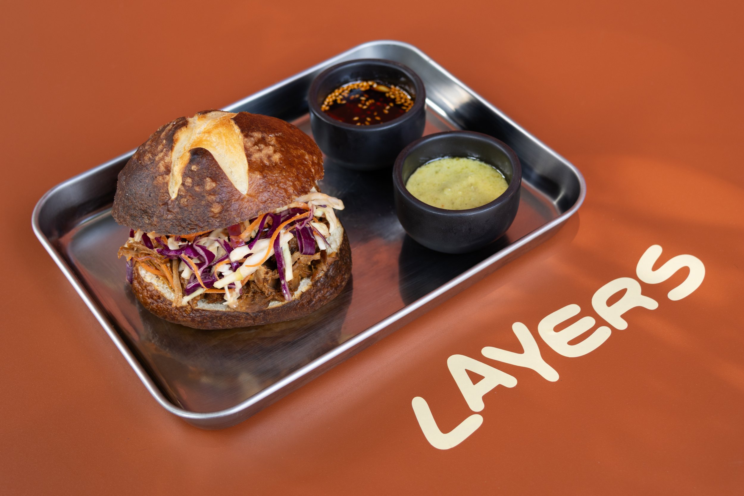 Una hamburguesa de pan frito con carne y ensalada en un plato con dos salsas negras. La mesa tiene la palabra 'LAYERS'.