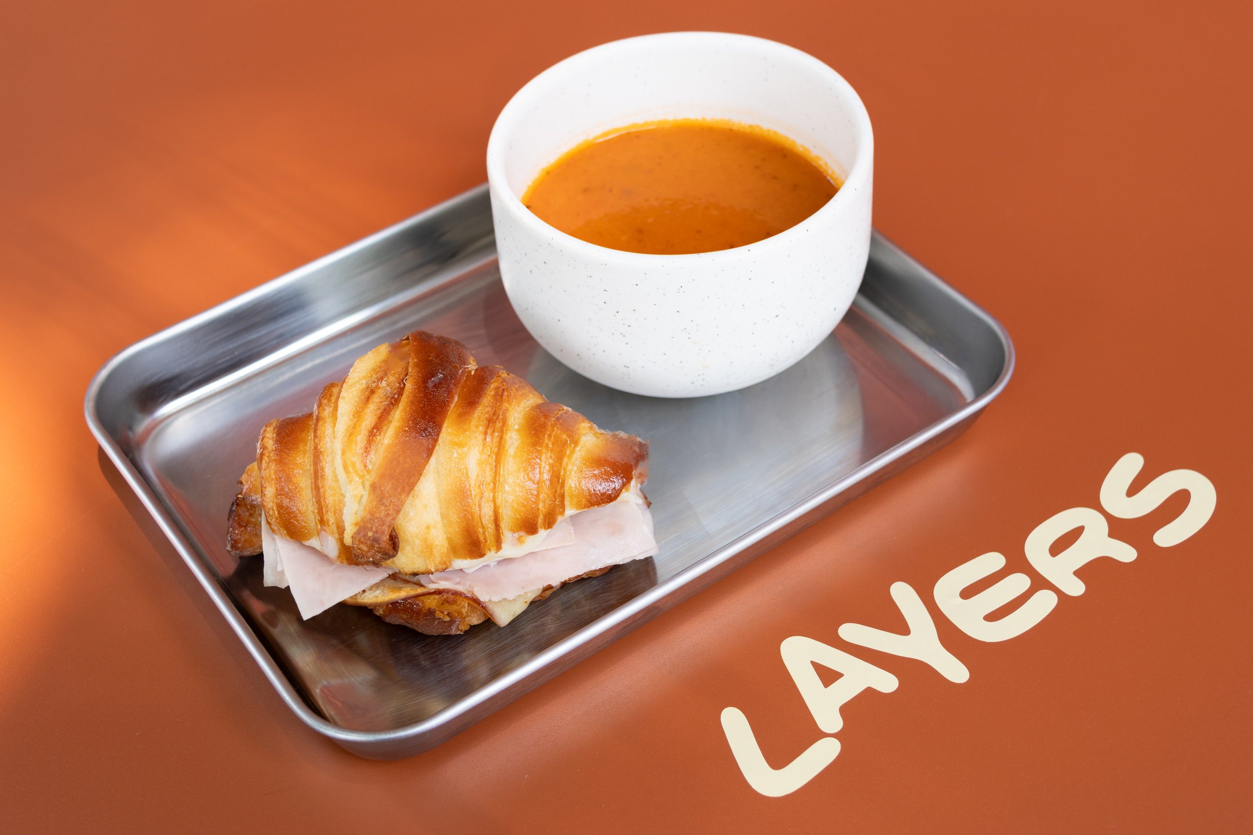 Un sándwich de jamón y queso con un croissant, acompañado de sopa en una taza blanca sobre una bandeja de metal en una mesa de color marrón.