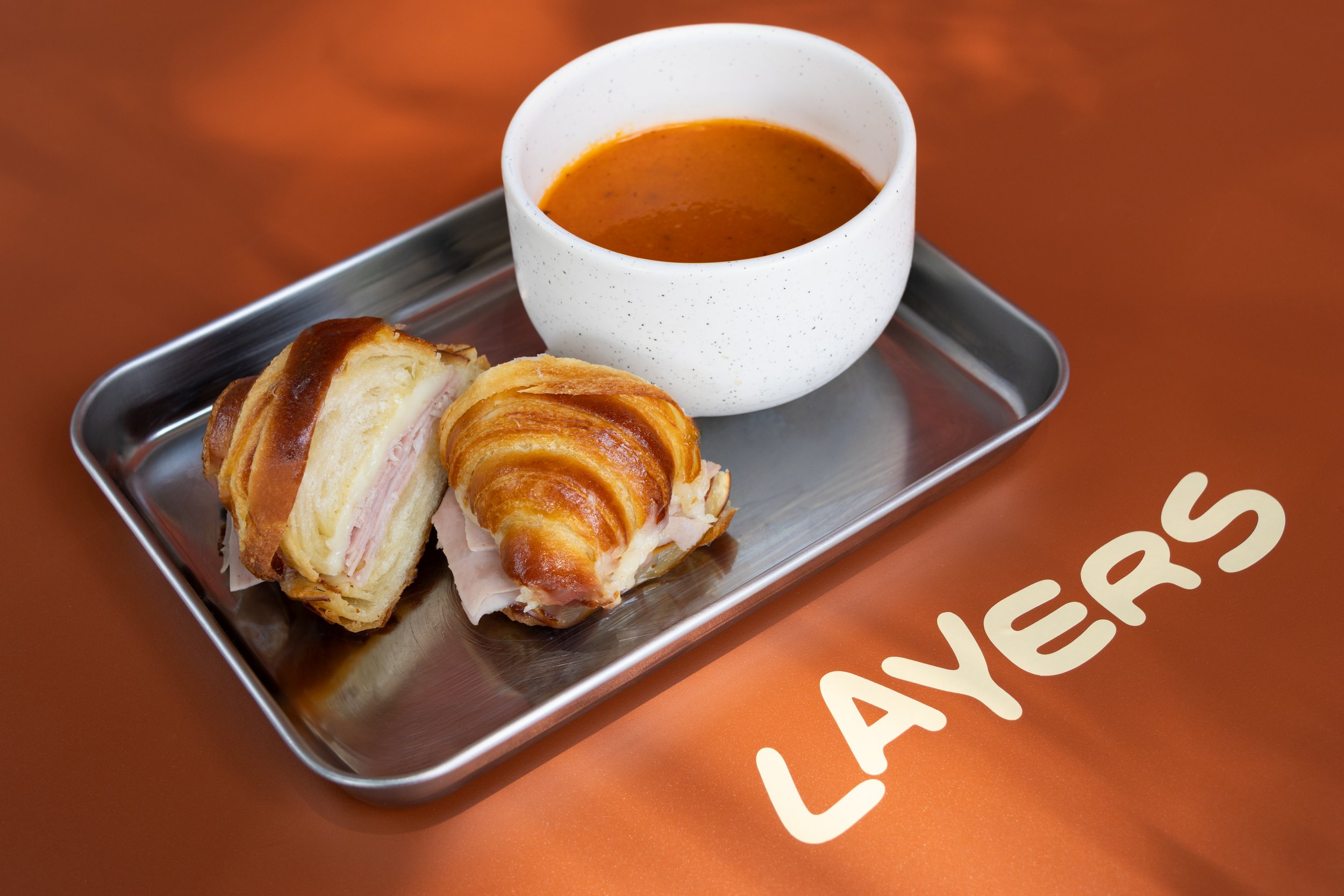 Sandwich de croissant con jamón y queso, acompañado de sopa en un tazón blanco, en una bandeja de metal sobre una mesa con la palabra "LAYERS" escrita en la superficie.