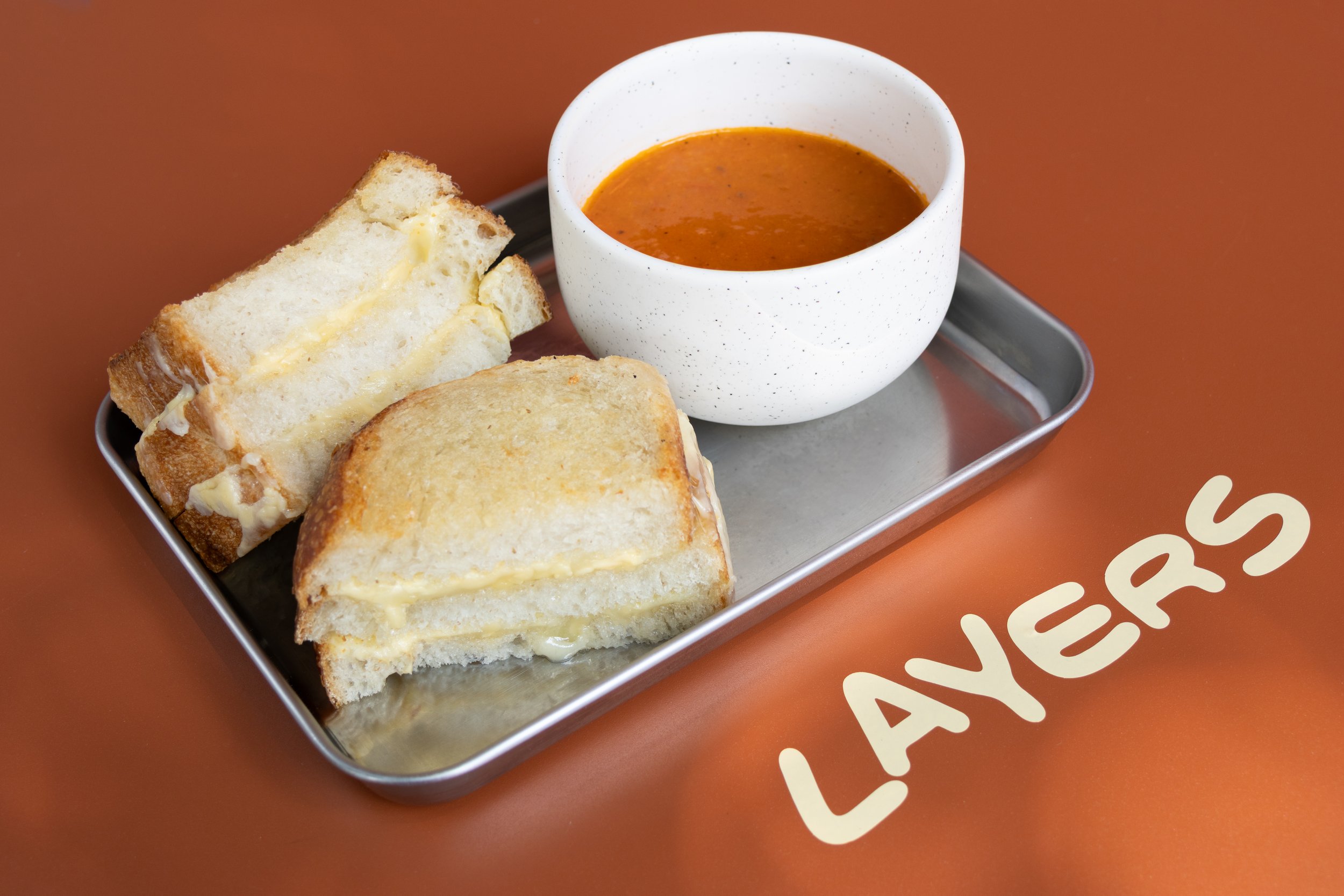 Sándwich de queso en pan tostado acompañado de sopa en plato y una taza blanca, sobre una mesa naranja con la palabra "Laivas"