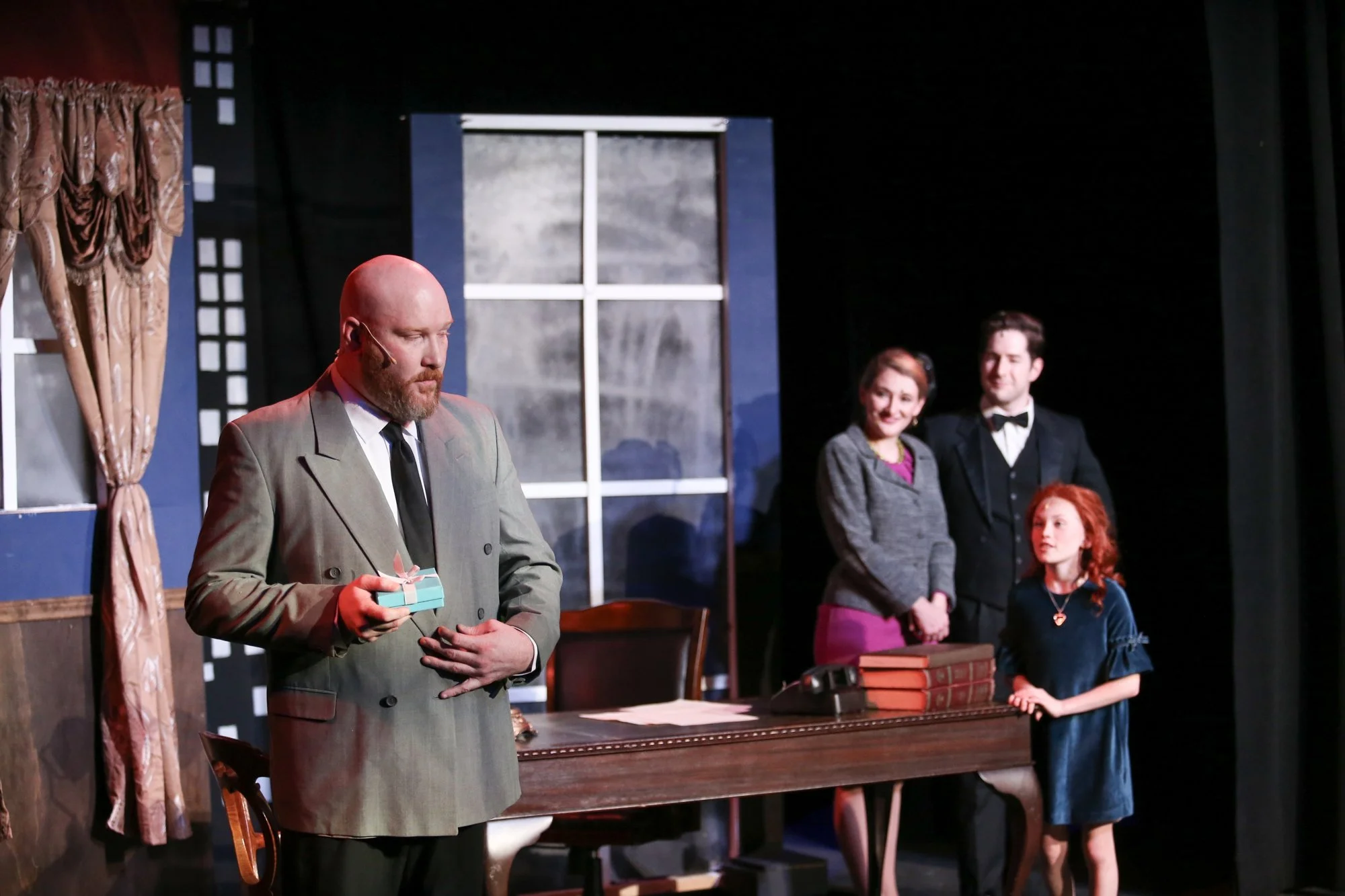 A scene from a theatrical performance featuring four actors on stage. The central actor, a bald man with a beard, is dressed in a suit and holding a small gift box. Three female actors and one young girl are standing in the background near a desk wit