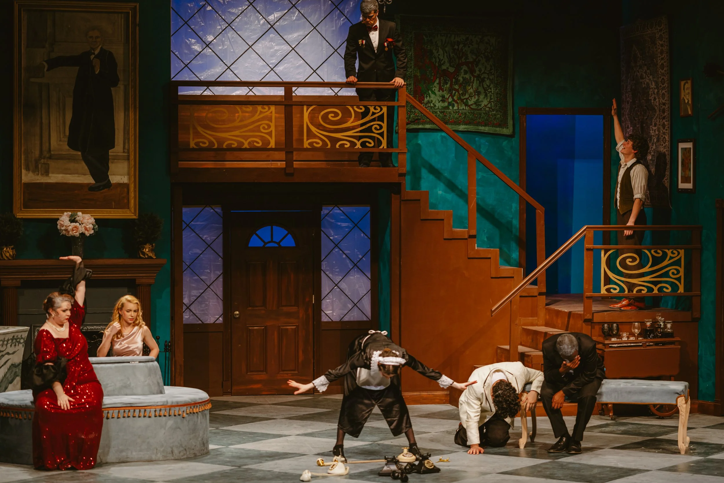 The scene depicts a theatrical play set in a luxurious, vintage-style interior with ornate furniture and decorations. Characters are engaged in an expressive scene, some on the ground and others standing, in period costumes. A large portrait hangs on