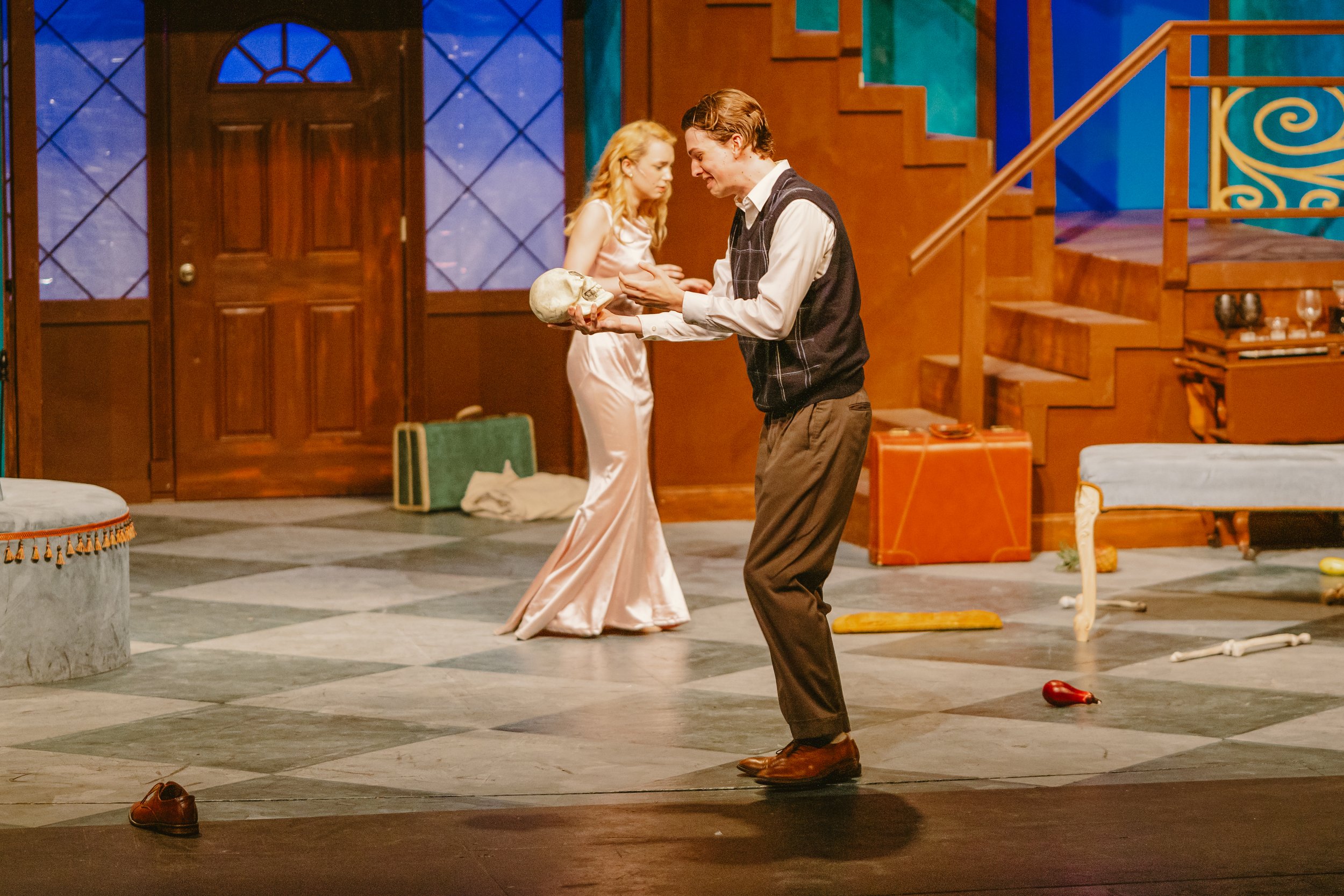 Two actors on stage with a wooden staircase and door in the background, one holding a skull, engaged in a theatrical scene.