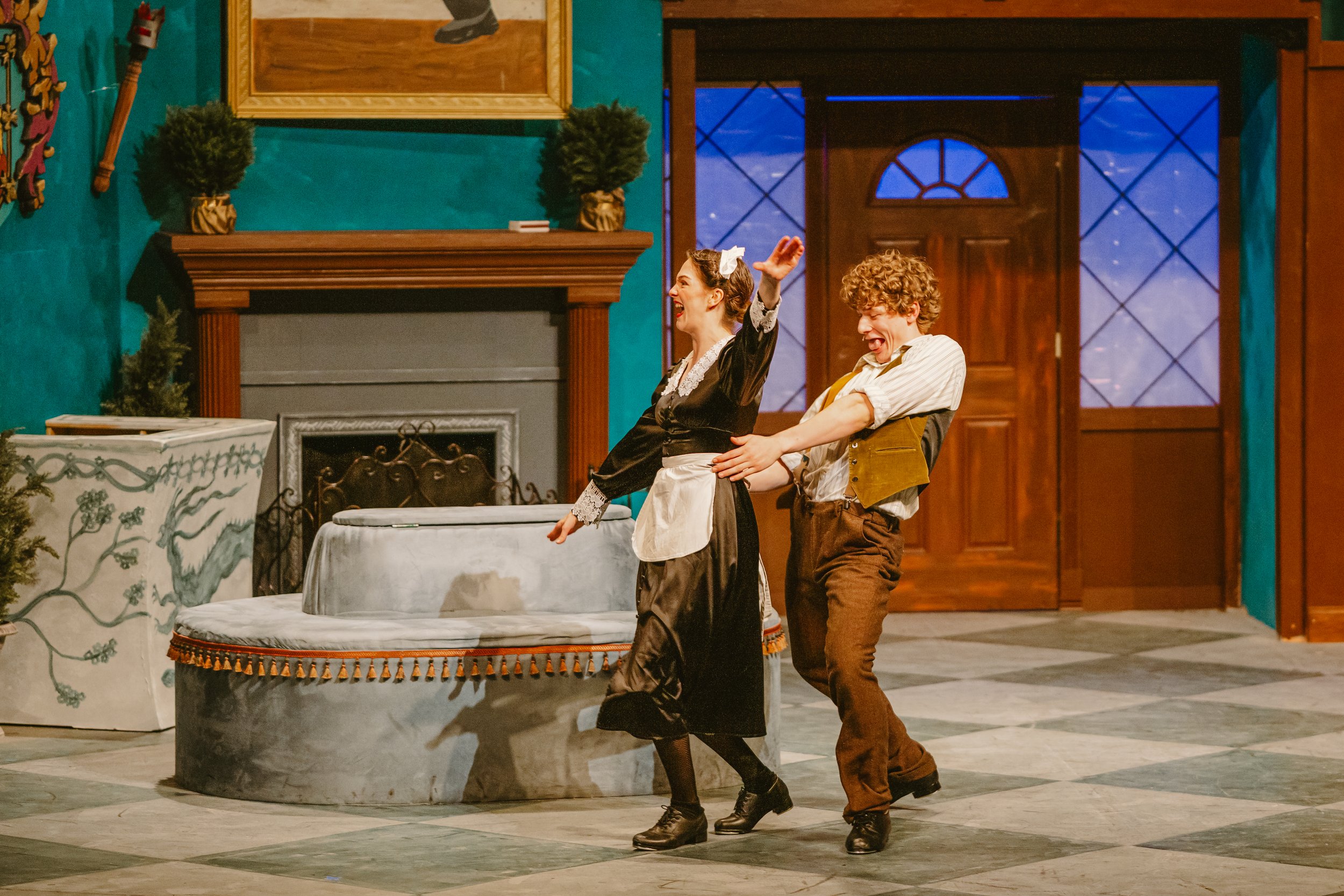 Two performers in vintage costumes on stage, one woman with a white bow in her hair and a black dress, and one young man with curly hair and a yellow vest, are playfully interacting near a round, decorated table in a set resembling a cozy, vintage li