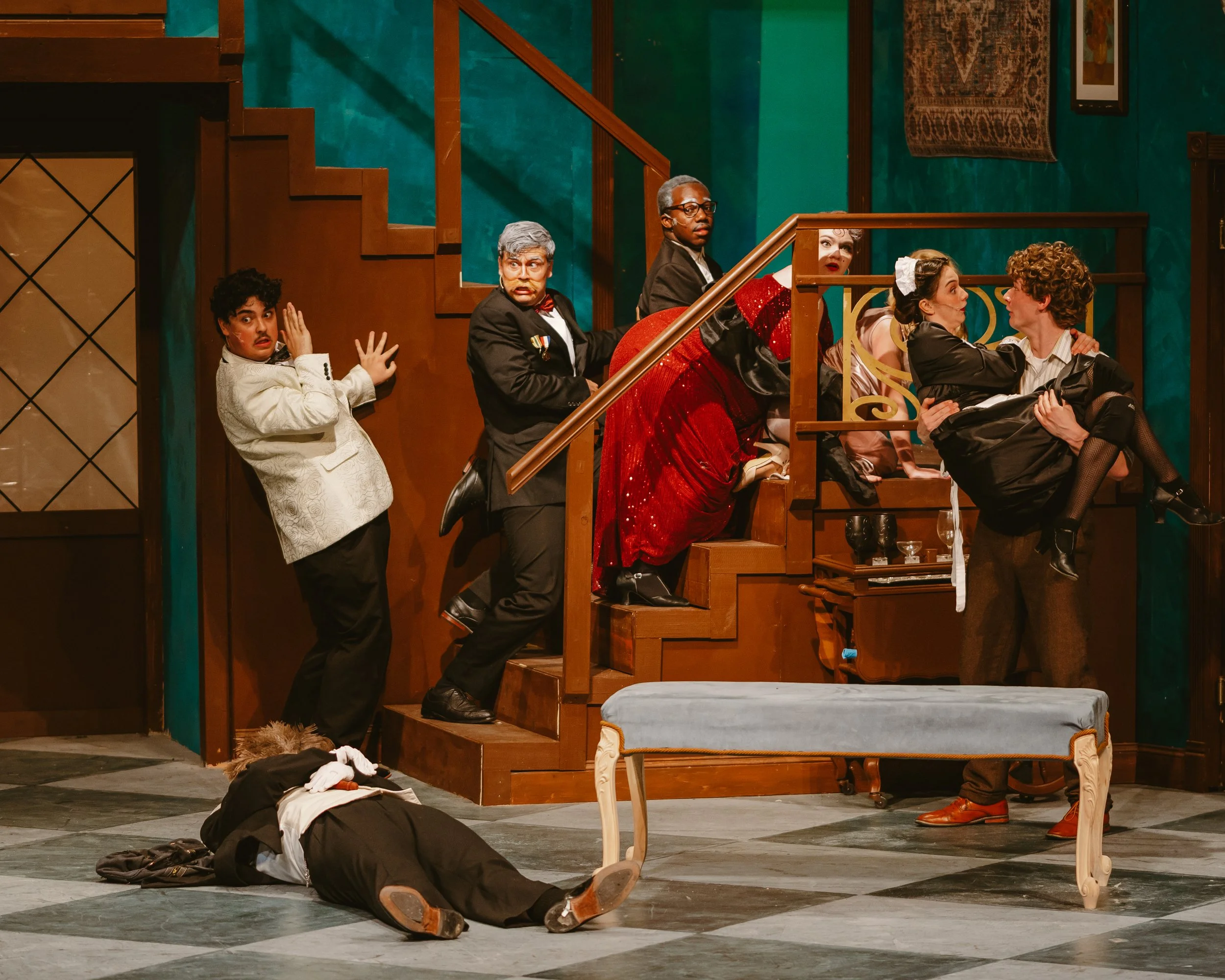 Stage scene with six actors in theatrical costumes portraying a lively or dramatic moment. One actor on the stairs appears surprised or scared, and two people on the right are engaged in an intense interaction, while others look on with various expre