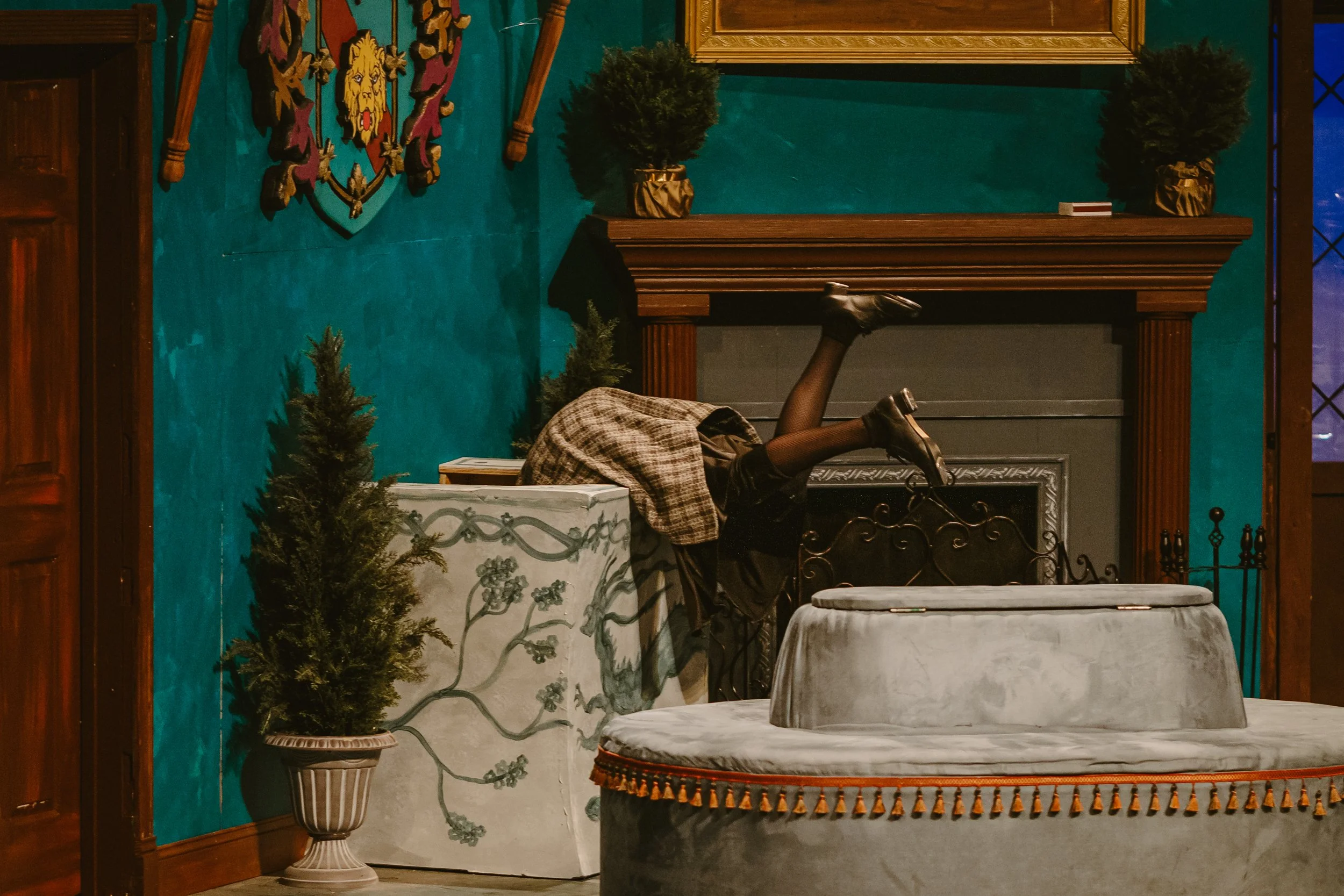A person lying face down on a table with their legs raised in the air and wearing black shoes, a plaid skirt, and black stockings. The scene is set indoors with decorative elements including a fireplace, two small trees in golden pots, a large framed